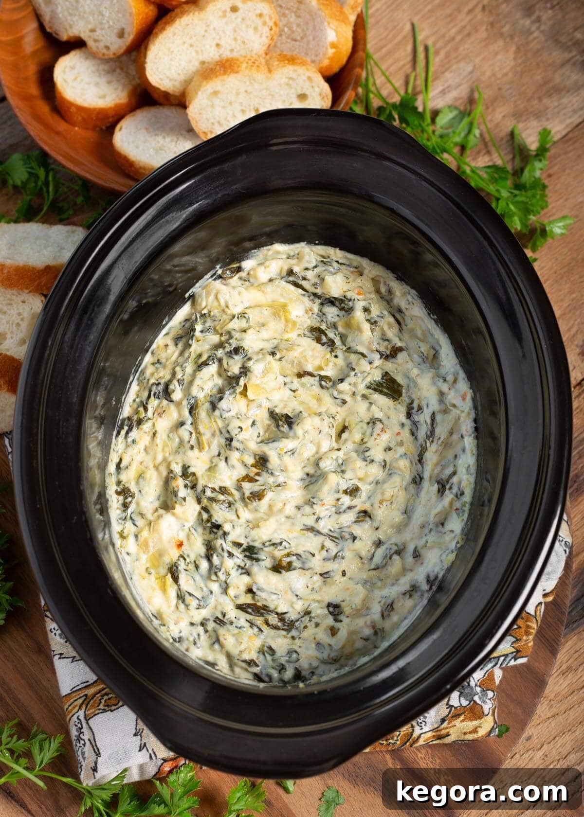 Slow Cooker Spinach Artichoke Dip in black crock with sliced baguette slices around it.