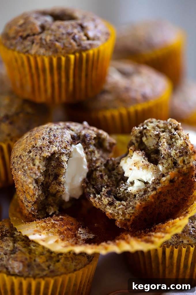 Close up of a perfectly split Chocolate Cream Cheese Muffin, proudly revealing its moist interior, flecks of rich chocolate, and the creamy, luscious orange-infused filling. Additional whole muffins are stacked softly blurred in the background, inviting indulgence.