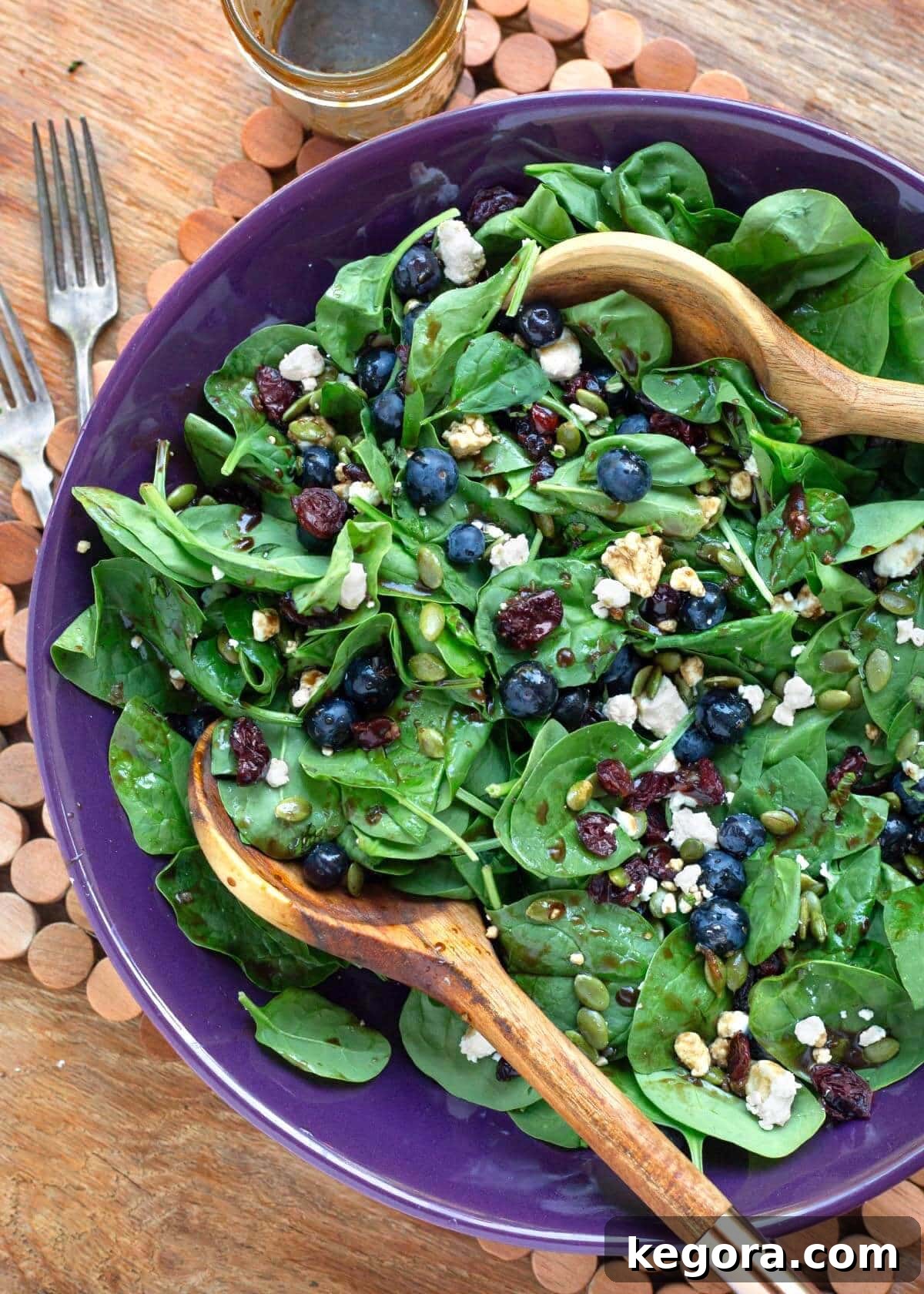 Fresh Blueberry Spinach Salad in a purple bowl, garnished with pumpkin seeds and dried cranberries, with wooden serving spoons.