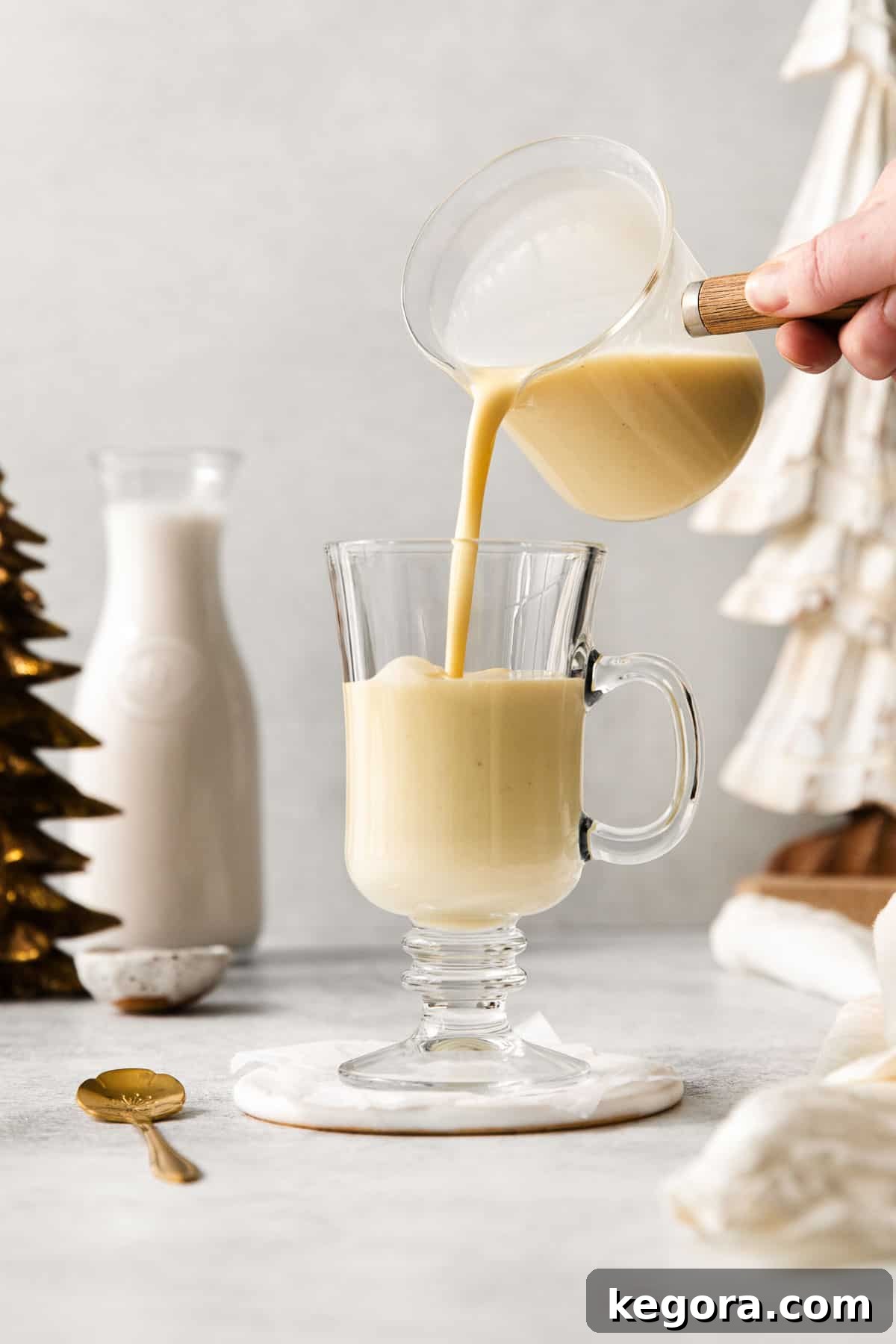 Spiced Dairy-Free Nog Delight 8 Thick, creamy homemade dairy-free eggnog being gracefully poured from a glass pitcher into a clear drinking glass, illustrating its rich texture.