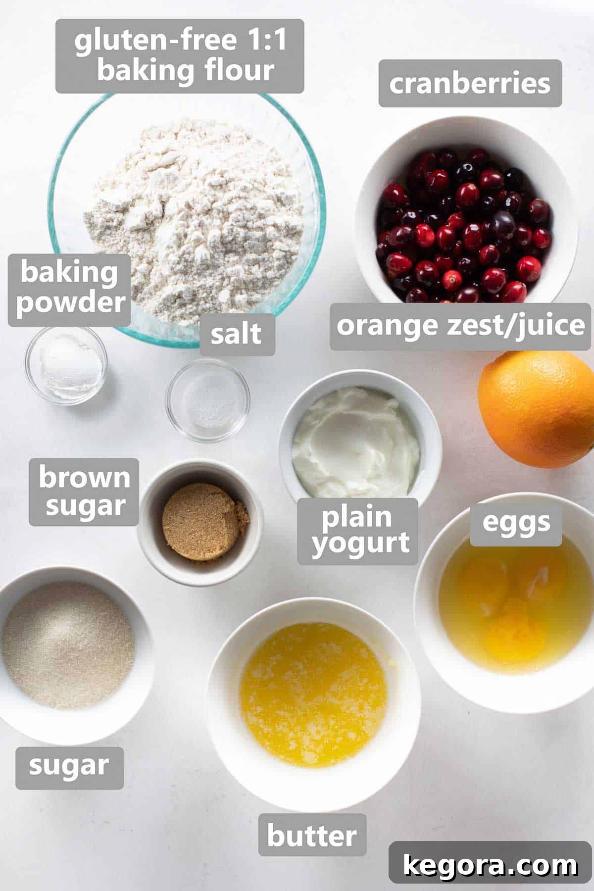Overhead view of various muffin ingredients neatly arranged in bowls on a pristine white background, showcasing flour, sugar, cranberries, and citrus.