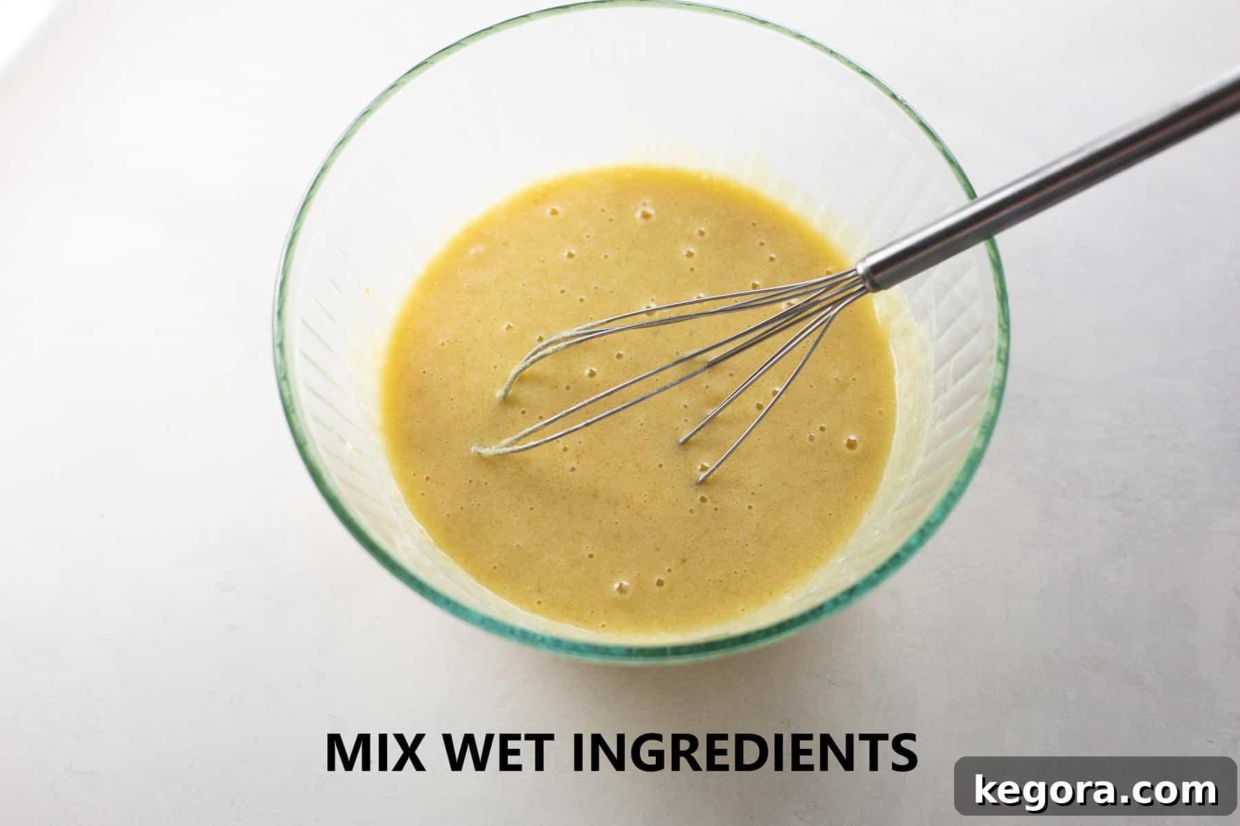 A large clear glass bowl holding a smooth, creamy mixture of wet muffin ingredients, including eggs, yogurt, and melted butter.