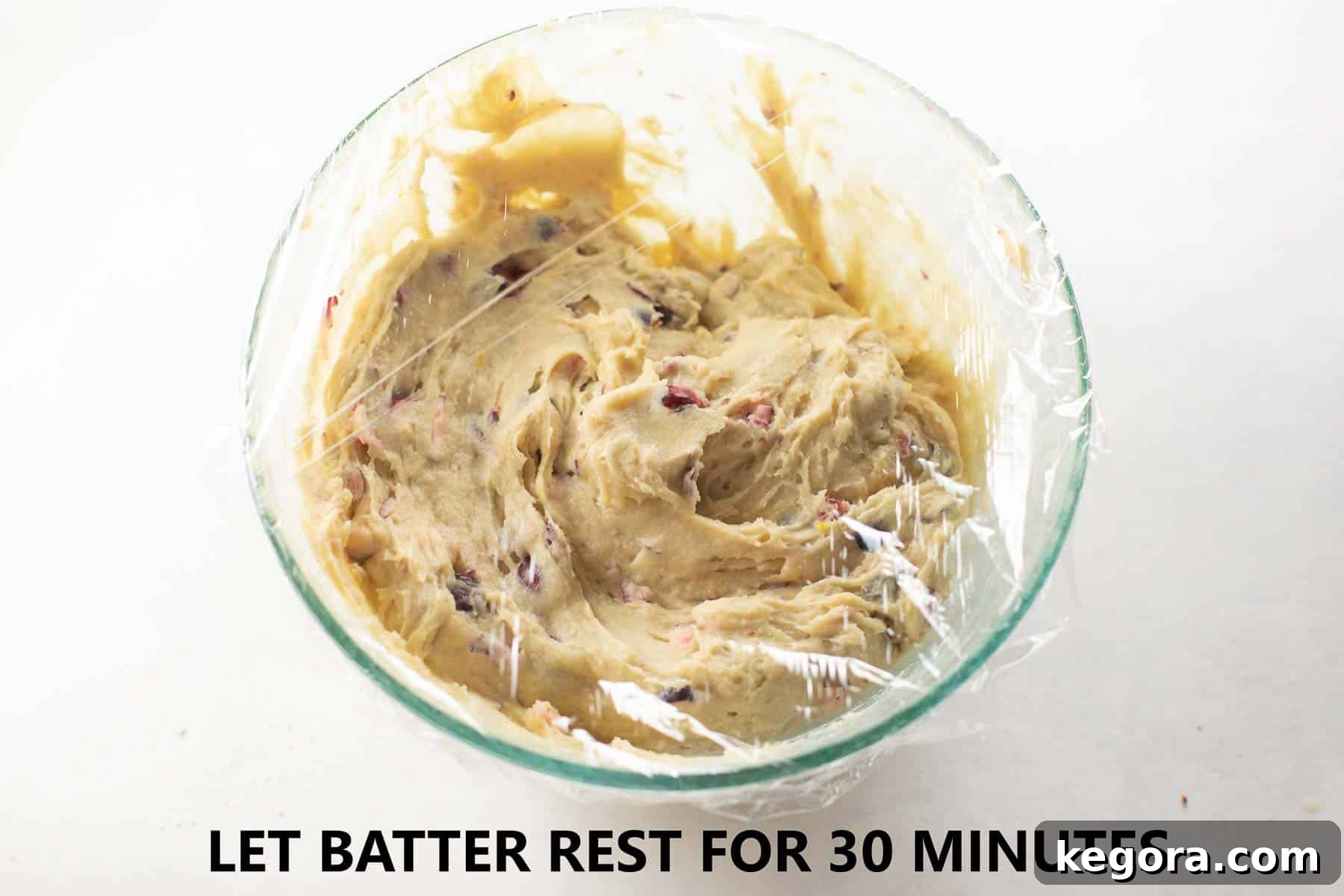 Muffin batter in a glass bowl, covered with plastic wrap, patiently resting on a kitchen counter.