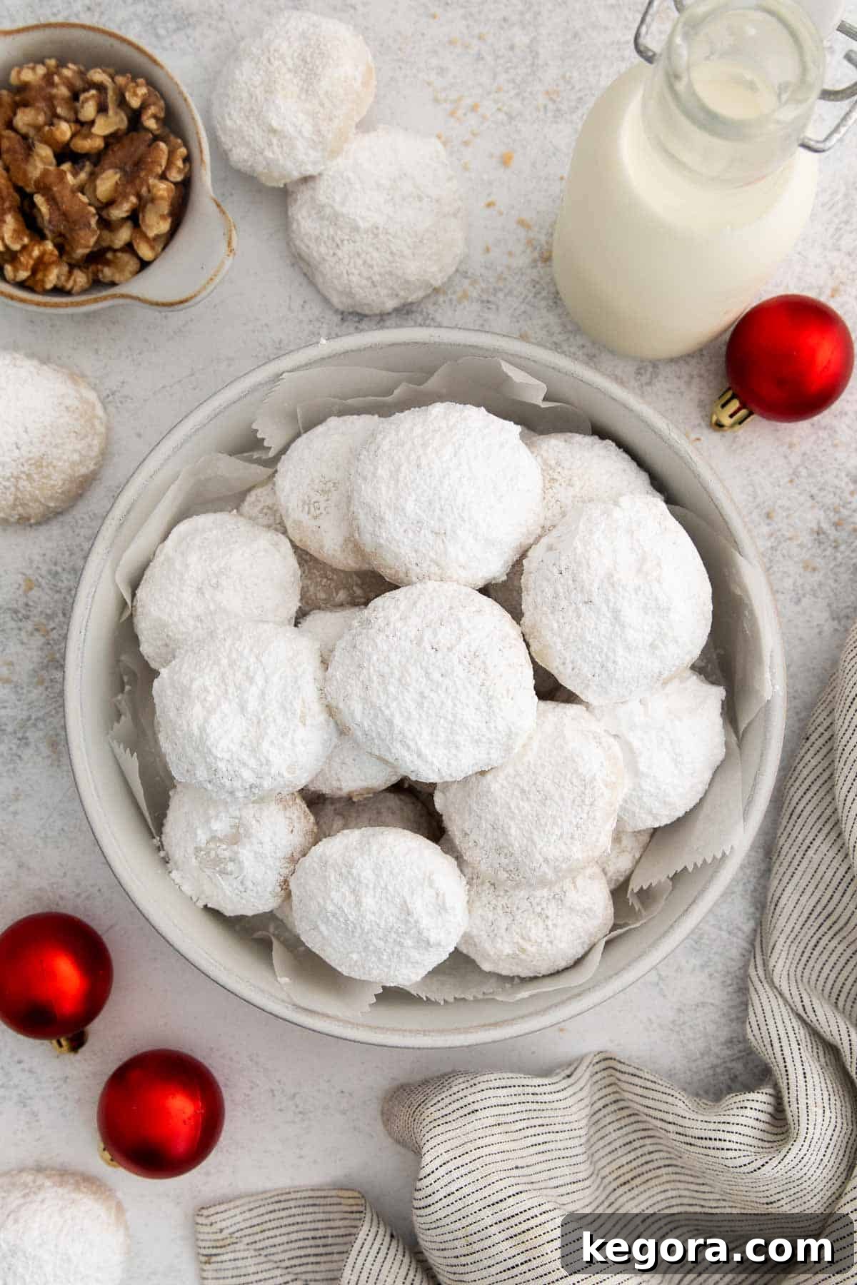 Dusted Dream Gluten-Free Snowballs 3 A bowl overflowing with freshly baked gluten-free snowball cookies, lightly dusted with powdered sugar, surrounded by festive red Christmas ornaments on a rustic counter. The image captures the inviting warmth and holiday spirit of homemade treats.