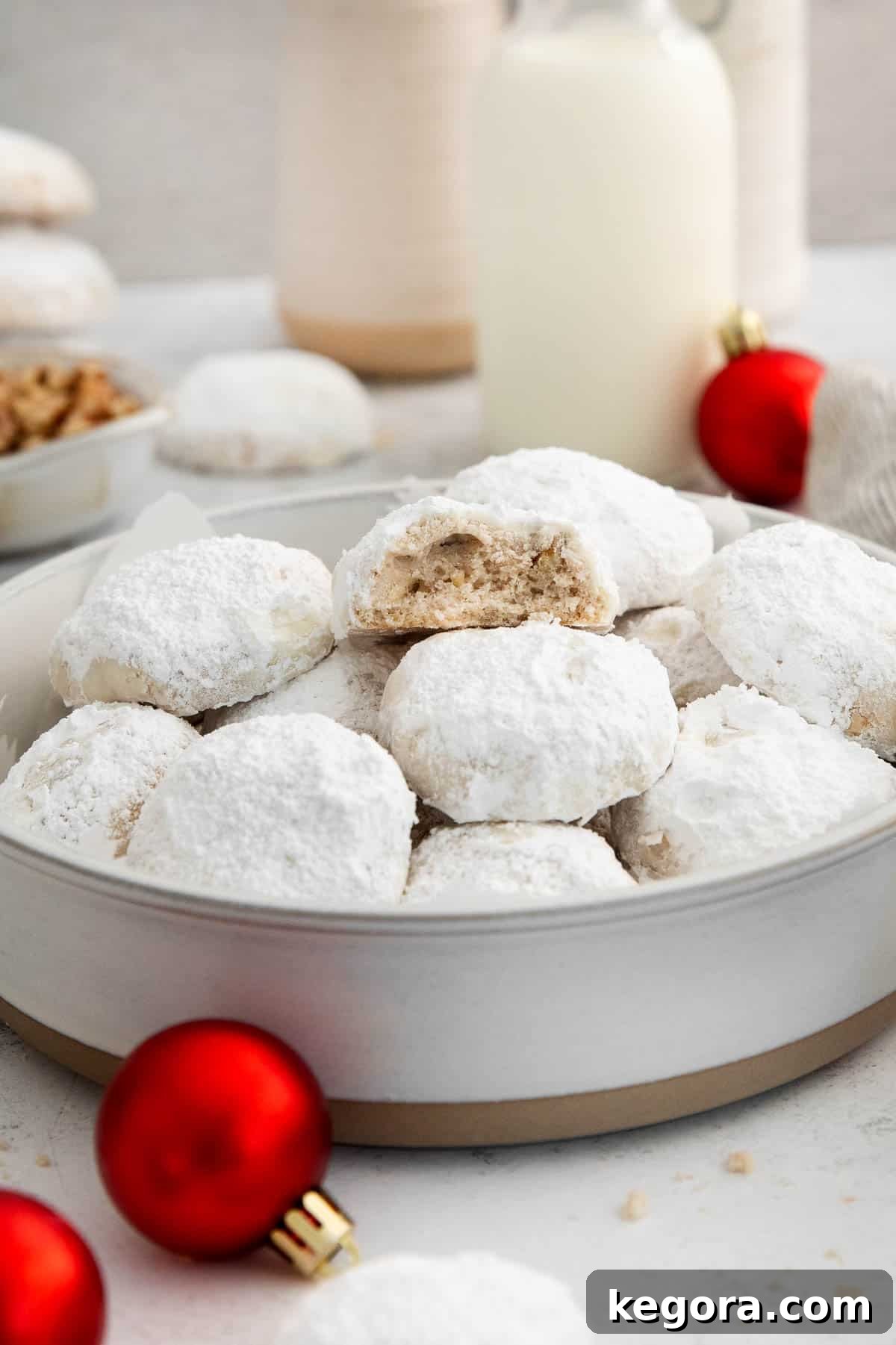 Dusted Dream Gluten-Free Snowballs 7 A close-up shot of a bowl full of gluten-free snowball cookies, with one cookie bitten into, revealing its tender interior, all surrounded by holiday decorations.