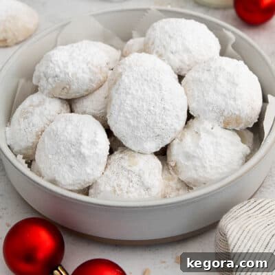 Dusted Dream Gluten-Free Snowballs 8 A close-up of a stack of snowball cookies in a bowl with red holiday ornaments, showcasing their snowy powdered sugar coating and festive appeal.