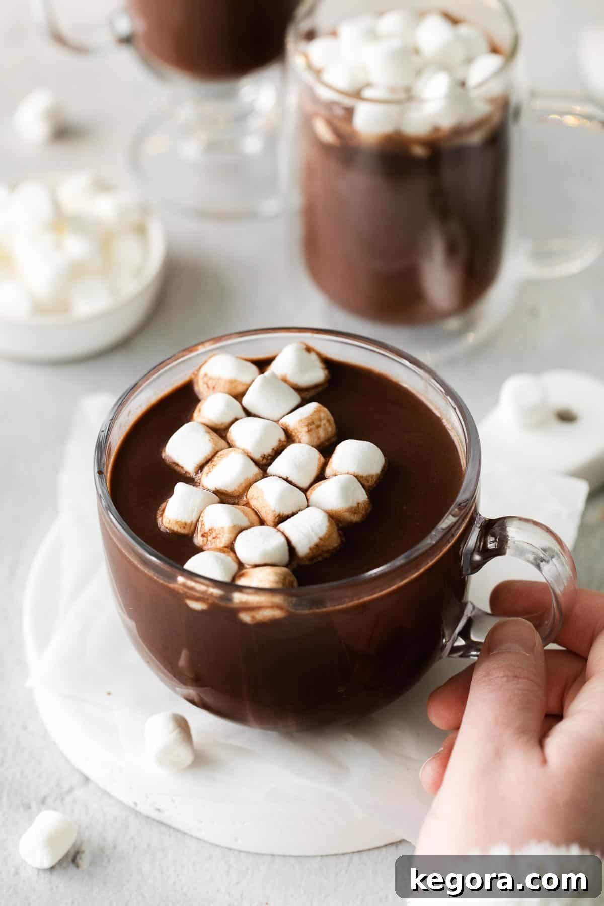 A hand holding a glass mug of hot chocolate with marshmallows on top, ready for a cozy moment