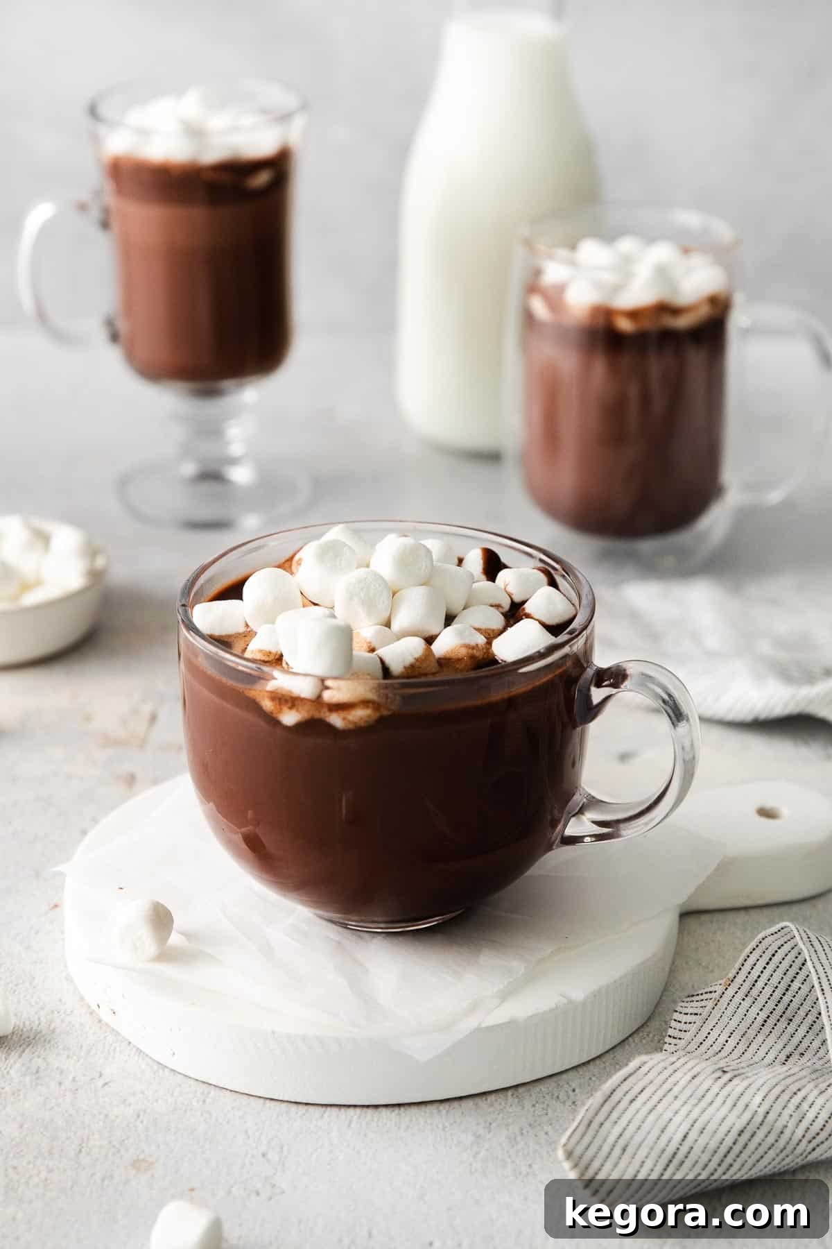 A glass mug of hot chocolate with marshmallows on top, and more mugs of hot chocolate in the background, inviting a moment of peace