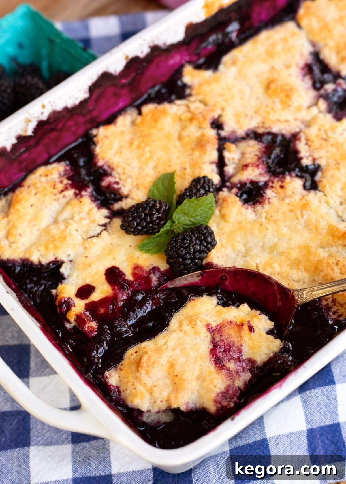 A large white baking dish filled with Homemade Blackberry Cobbler, with a serving spoon resting inside, showcasing the deep berry color and golden biscuit topping.