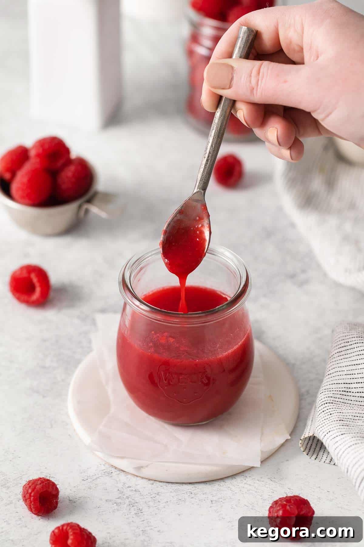 Vibrant Raspberry Drizzle 8 A hand gracefully dipping a silver spoon into a glass jar brimming with luscious raspberry coulis, highlighting its smooth texture.
