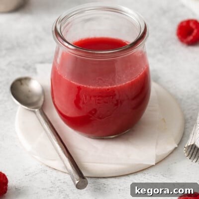 A glass jar filled with vibrant raspberry coulis, with a serving spoon next to it, showcasing its appealing texture.
