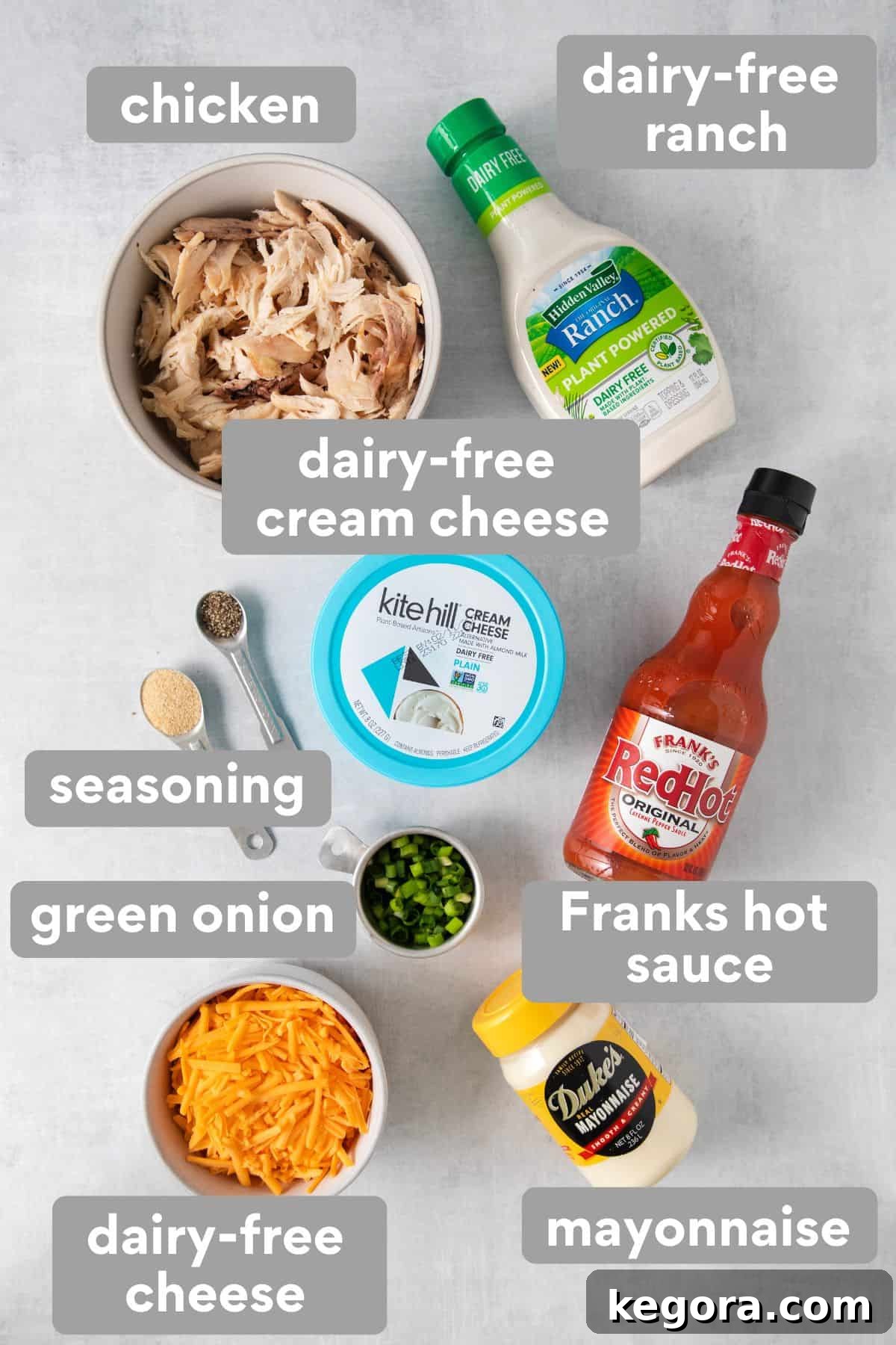 All the essential dairy-free buffalo chicken dip ingredients laid out on a kitchen countertop: shredded chicken, dairy-free cream cheese, dairy-free ranch, Frank's RedHot sauce, dairy-free mayonnaise, dairy-free cheddar shreds, and fresh green onions, alongside garlic powder and black pepper.