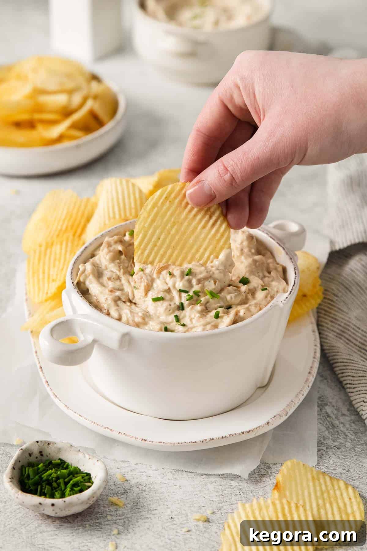 Ultimate Caramelized Onion Dip 8 A hand gracefully dipping a crinkle-cut potato chip into a bowl of perfectly creamy caramelized onion dip, beautifully showcasing the inviting and luxurious texture of the dip, ready for enjoyment.