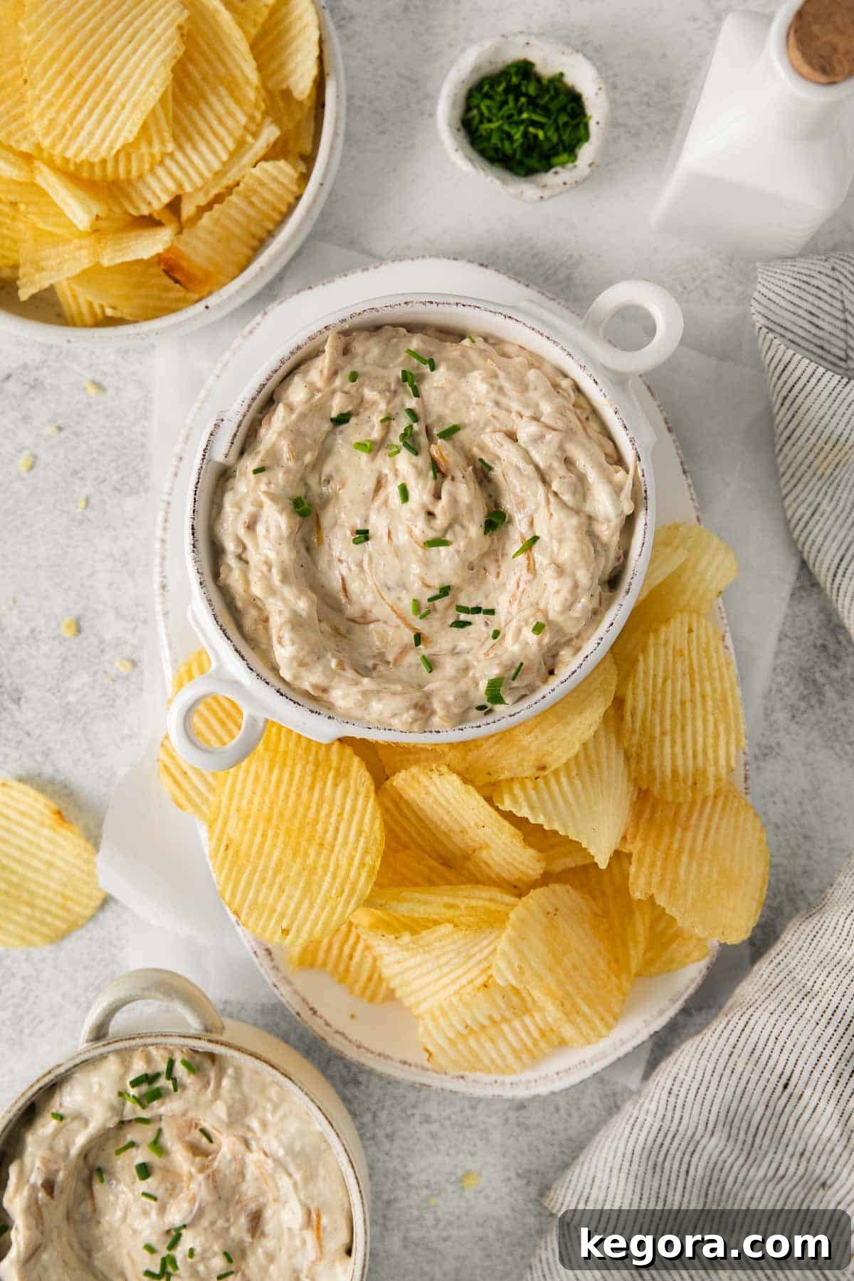 Ultimate Caramelized Onion Dip 9 Two enticing bowls of homemade caramelized onion dip are displayed, one with a serving spoon nestled within, and the other elegantly surrounded by an assortment of crispy potato chips, collectively highlighting its undeniable appeal as the ultimate party snack.