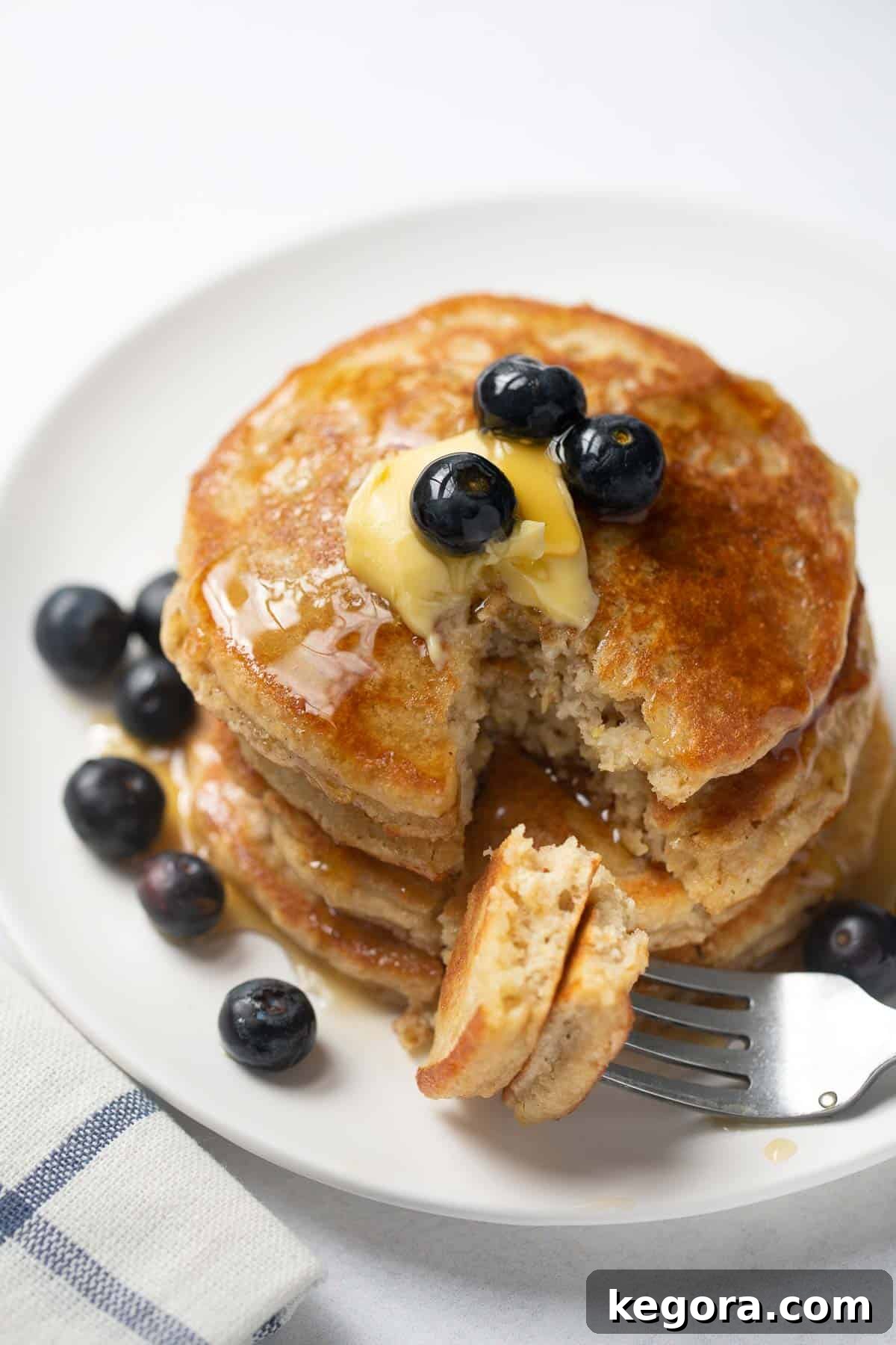 Gluten-Free Pancakes Ready to Eat Tall stack of gluten-free pancakes on a white plate, garnished with blueberries and drizzled with maple syrup, with a fork taking a bite