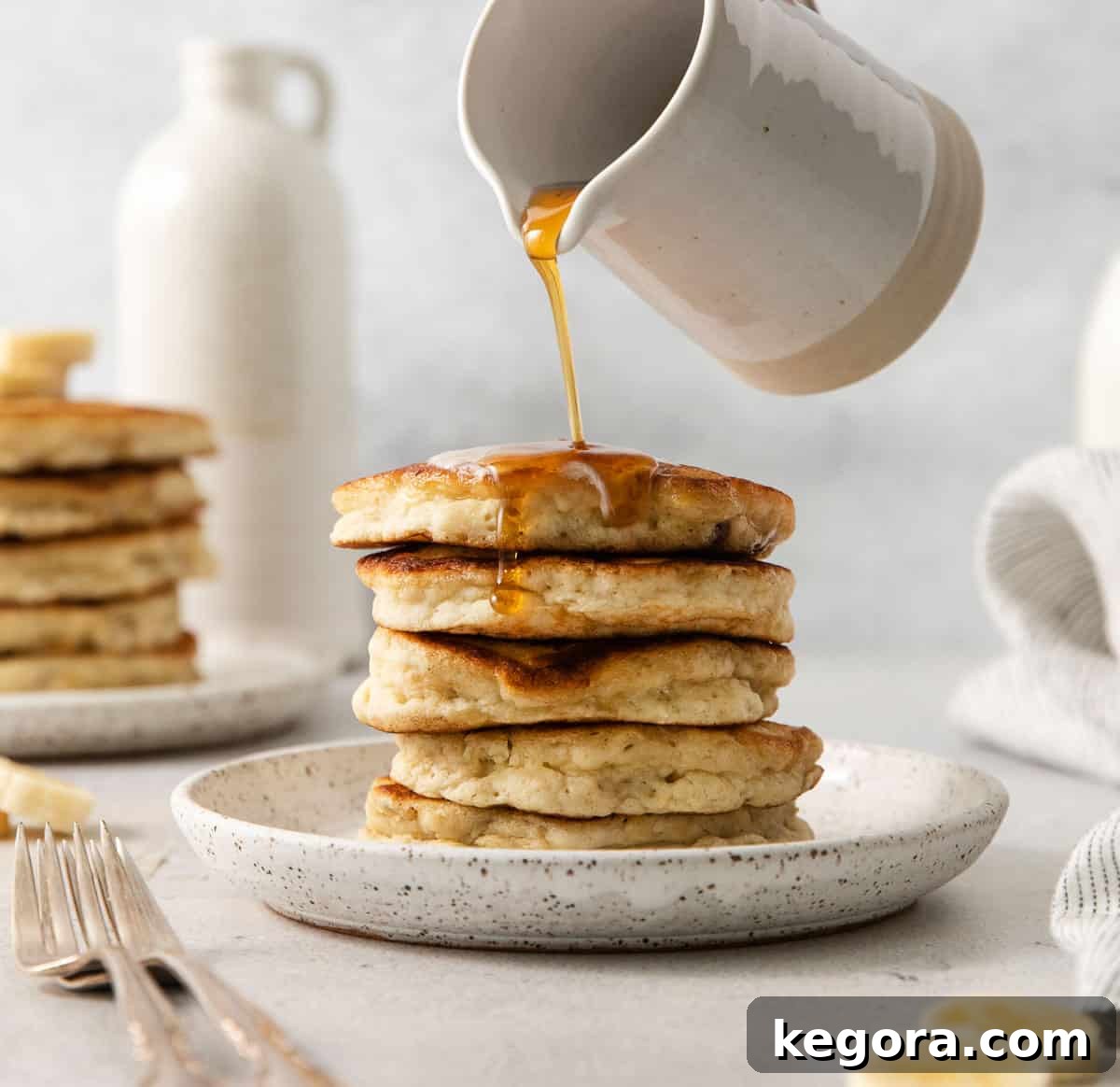 Naturally Sweet Gluten-Free Banana Hotcakes 9 Gluten-free banana pancakes stacked up on a plate with a hand pouring syrup over them, highlighting their appealing texture.