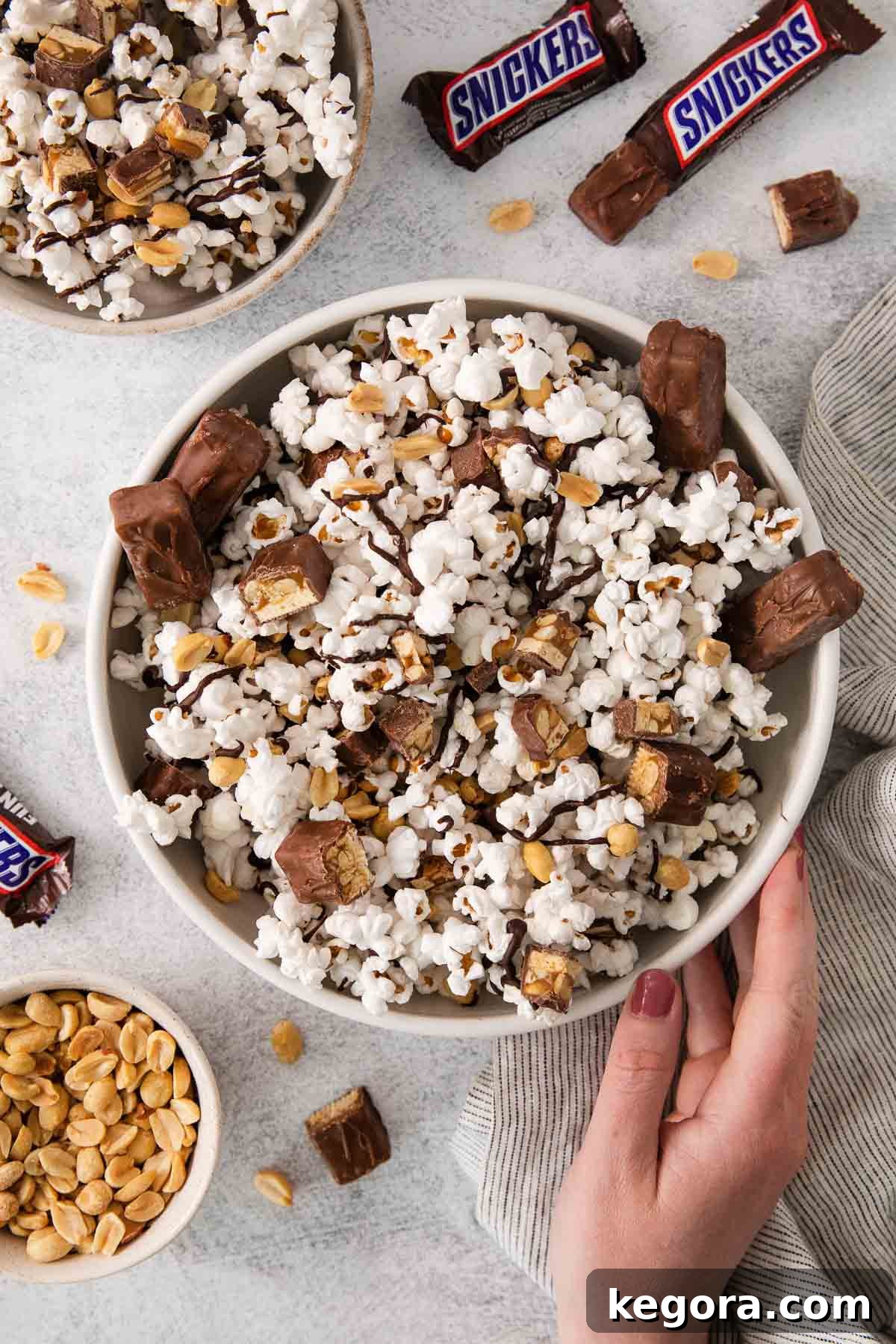 SnickerPop Crunch 2 A large bowl of homemade Snickers popcorn, perfectly drizzled with chocolate and mixed with chopped Snickers and peanuts, ready to be enjoyed. A hand reaches in to grab a piece, highlighting its inviting texture.