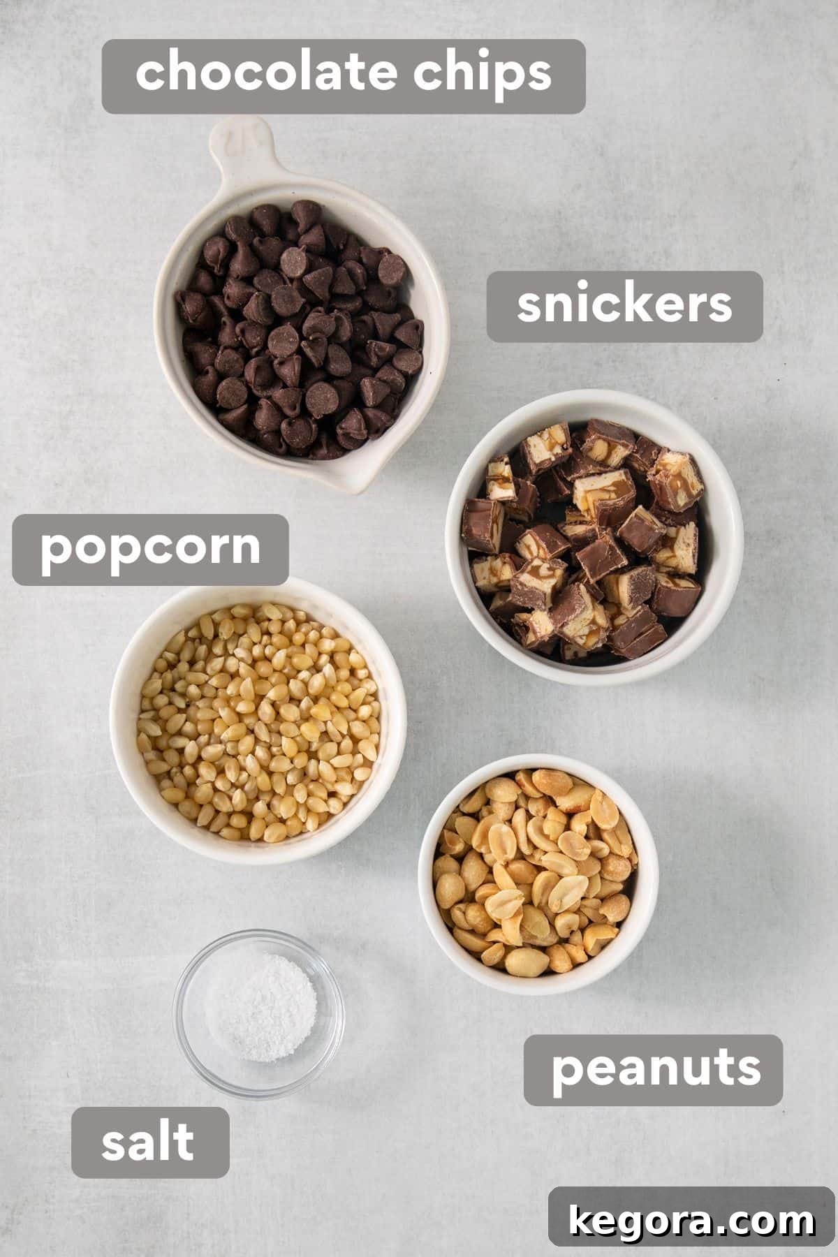 SnickerPop Crunch 3 Arrangement of Snickers popcorn ingredients including chocolate chips, whole Snickers bars, raw popcorn kernels, and roasted peanuts laid out on a clean countertop, ready for preparation.