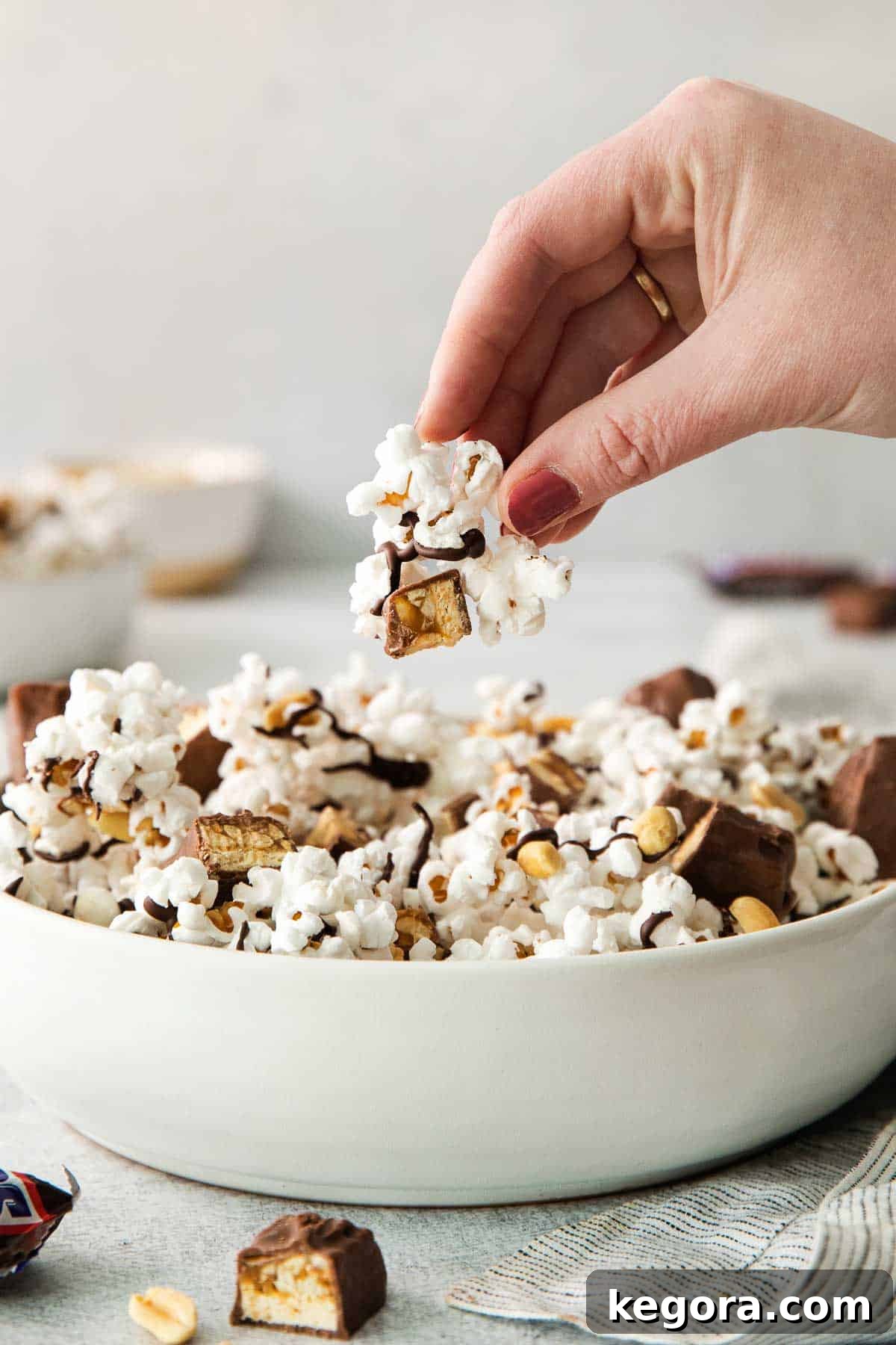 SnickerPop Crunch 9 Close-up of Snickers popcorn in a bowl with a hand reaching in to pick up a piece, emphasizing the texture and deliciousness.