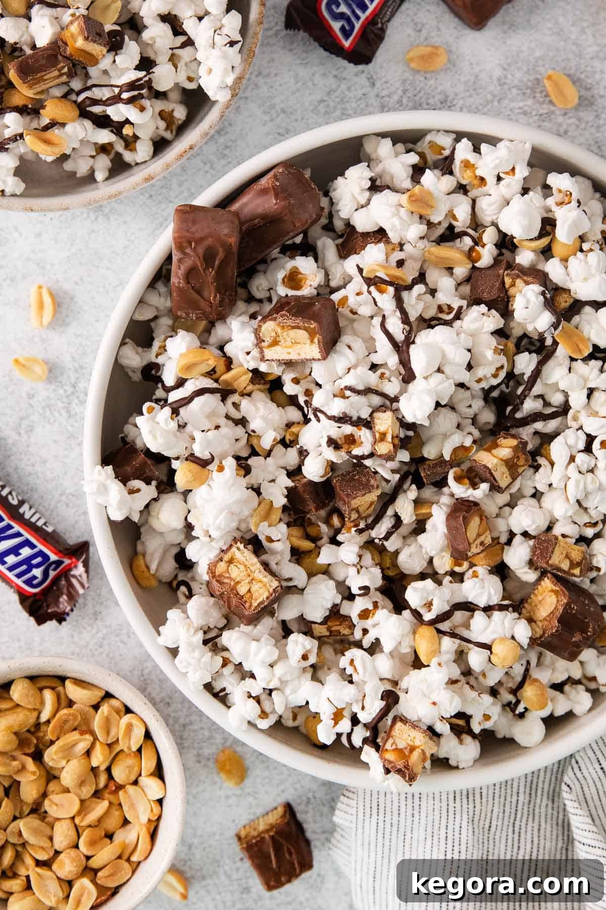 SnickerPop Crunch 10 A bowl brimming with Snickers popcorn, accompanied by a whole Snickers bar and a scattering of peanuts on the side, highlighting the inspiration for the snack.