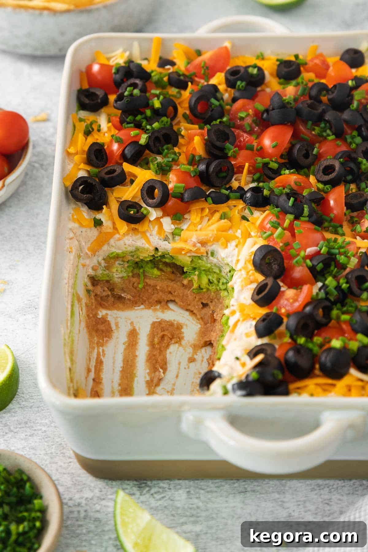 A vibrant 7-layer taco dip served in a clear baking dish, showcasing distinct layers of beans, guacamole, sour cream, and fresh toppings. A portion has been scooped out, ready for enjoyment.