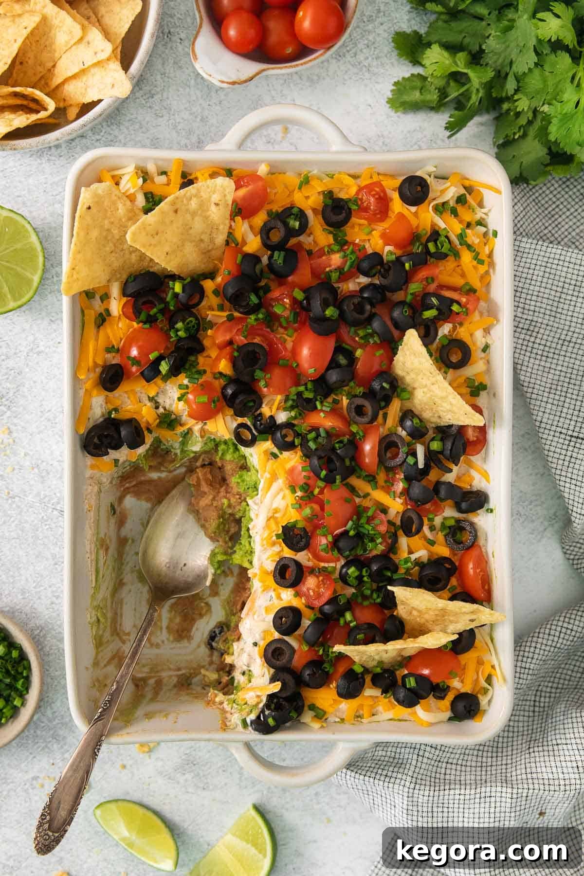 An inviting overhead shot of a freshly made 7-layer taco dip in a clear baking dish, with a serving spoon having scooped out a generous portion, emphasizing its readiness for guests.