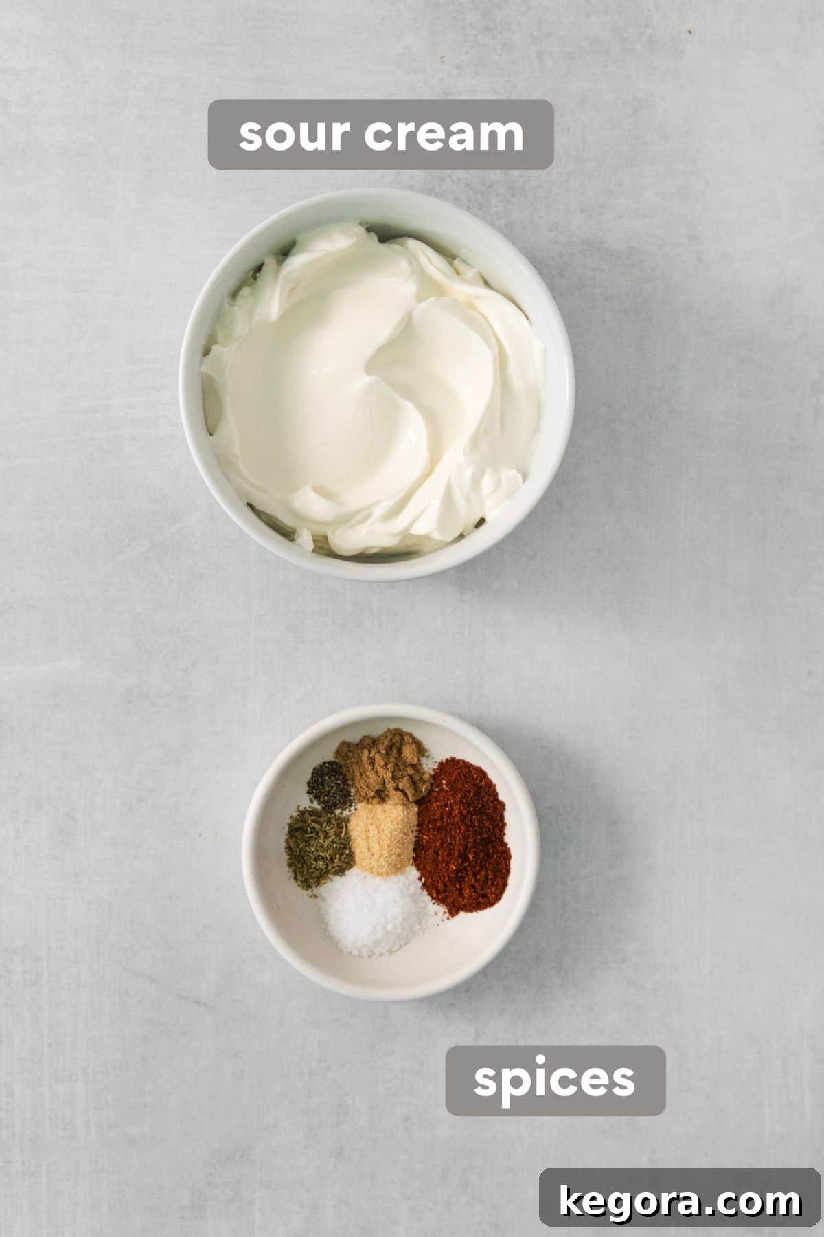 A bowl of creamy sour cream next to a small dish of ground spices, including chili powder and cumin, for seasoning the sour cream layer.