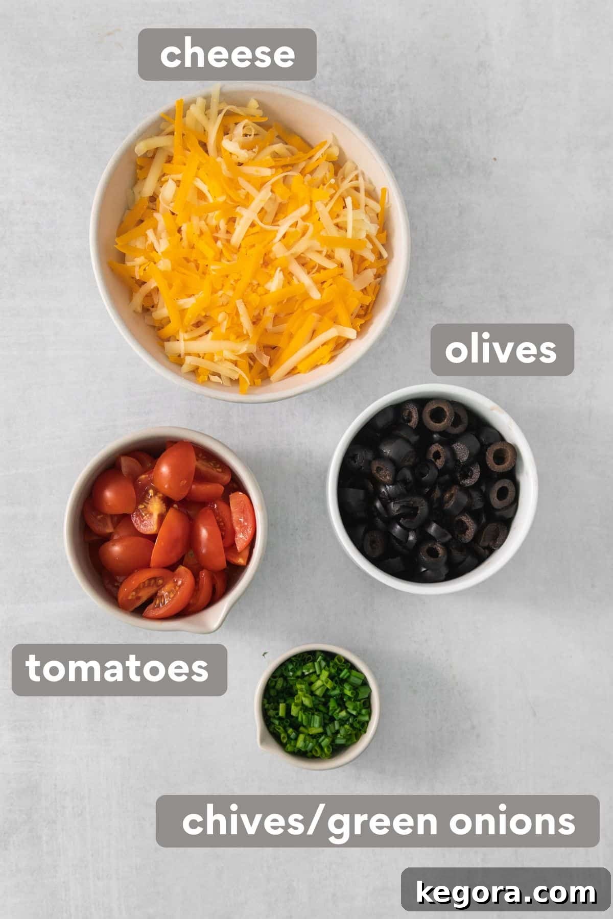 A selection of fresh toppings for 7-layer taco dip: shredded cheese, diced tomatoes, sliced black olives, and chopped green onions, arranged on a kitchen countertop.