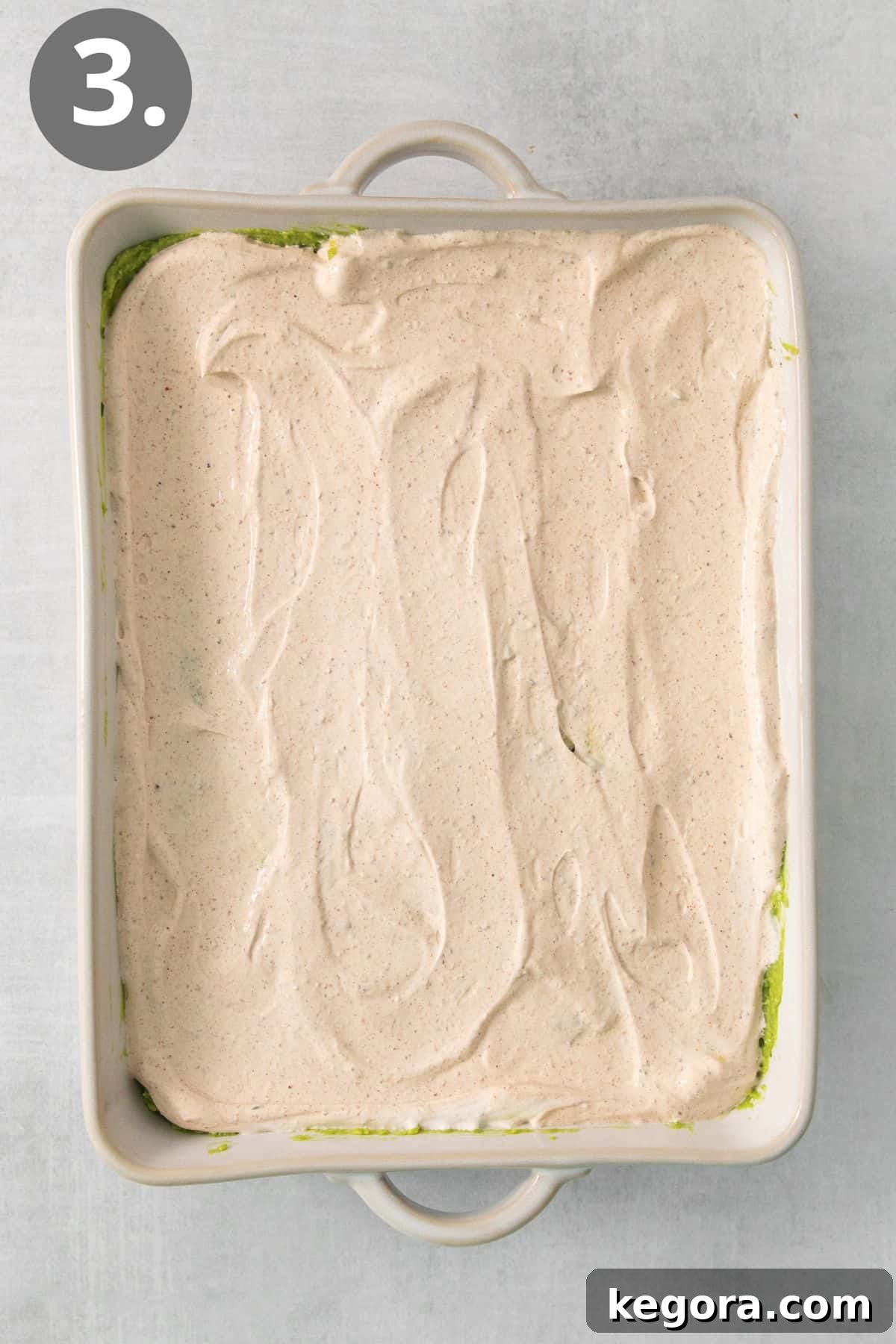 The creamy, seasoned sour cream layer meticulously spread over the guacamole and refried beans in a baking dish, ensuring full coverage.