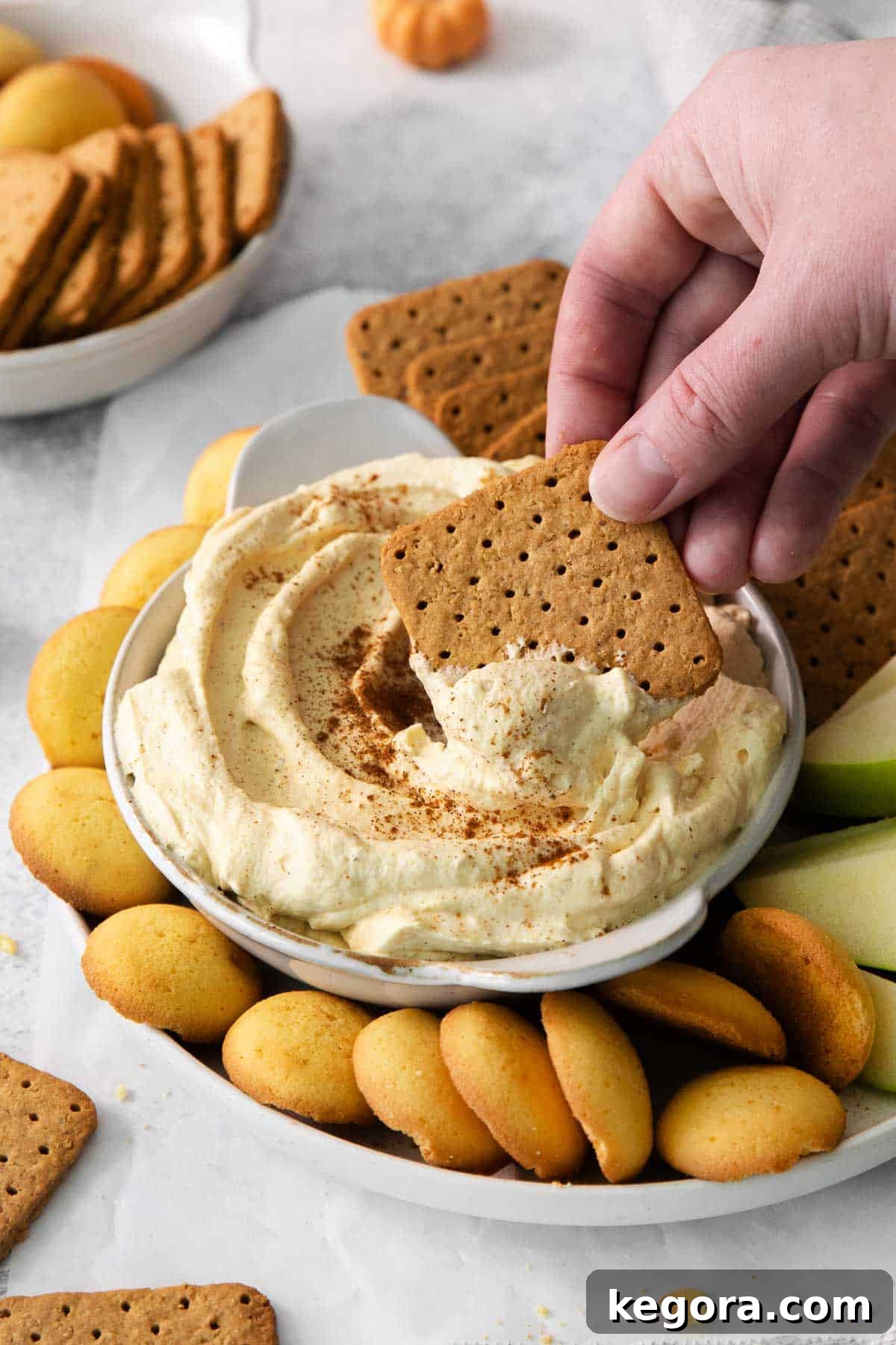 A hand dipping a gluten-free graham cracker into a bowl of creamy pumpkin cheesecake dip, surrounded by other dippers.