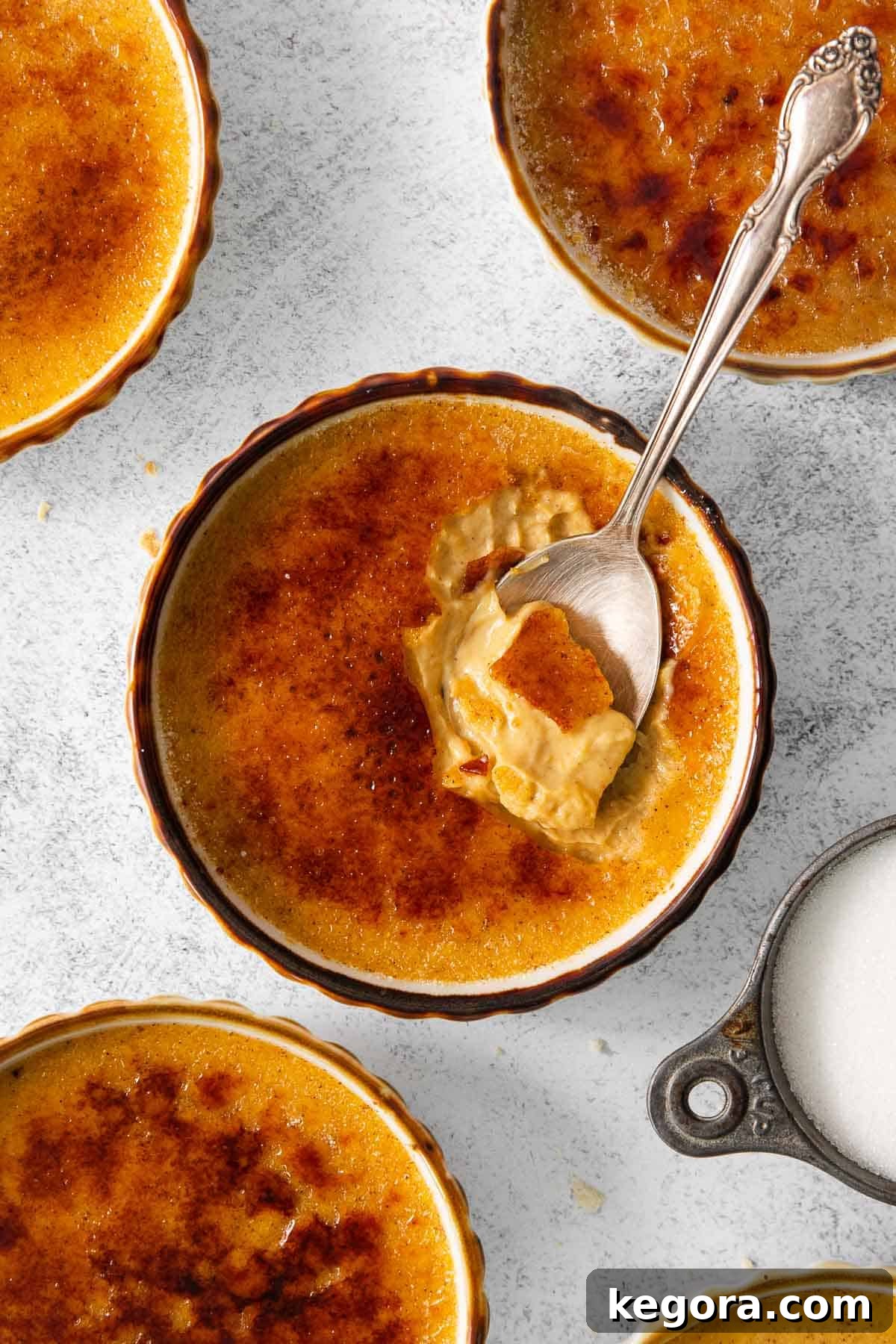 Velvet Pumpkin Creme Brulee 3 Pumpkin creme brulee in ramekins on a countertop, with a spoon in the ramekin, ready to be enjoyed.