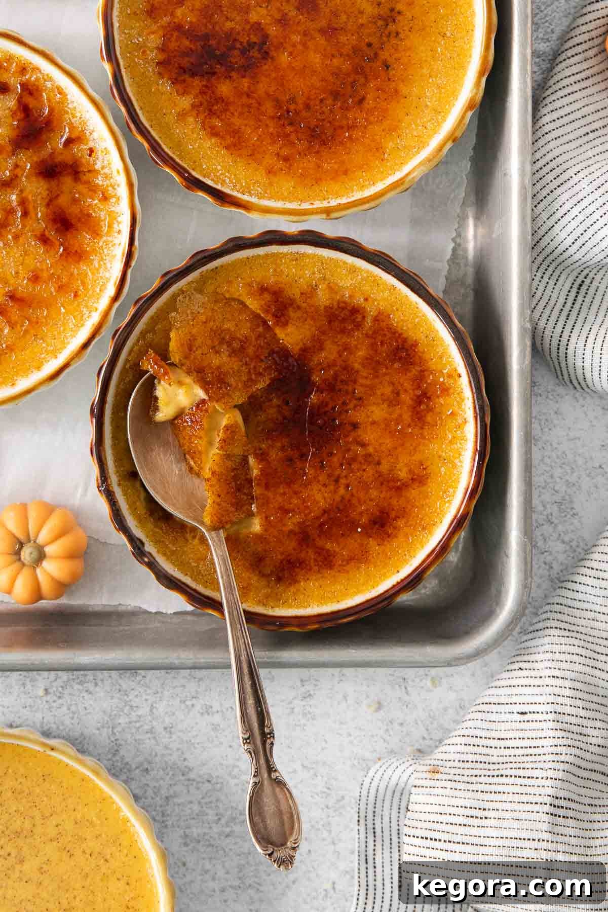 Velvet Pumpkin Creme Brulee 9 Close-up of a pumpkin creme brûlée in a ramekin on a baking tray with a spoon resting in it, ready to be eaten.