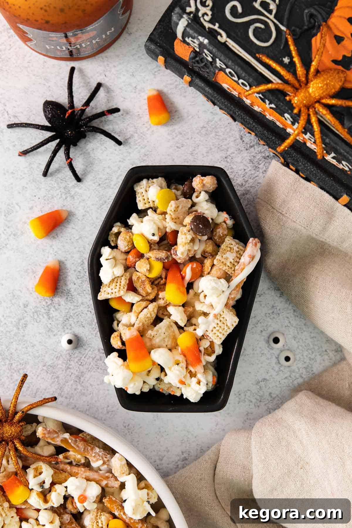 Monster Munch Mix 9 A festive Halloween bowl filled with snack mix, surrounded by spooky decorations like small pumpkins and spiders.