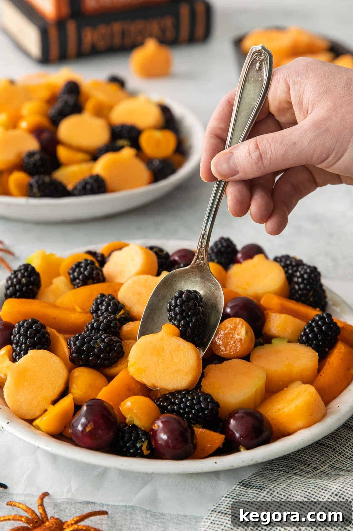Monster Mash Fruit Salad 8 A hand holding a serving spoon, about to scoop a portion from the large bowl of Halloween fruit salad, highlighting its freshness and appeal.