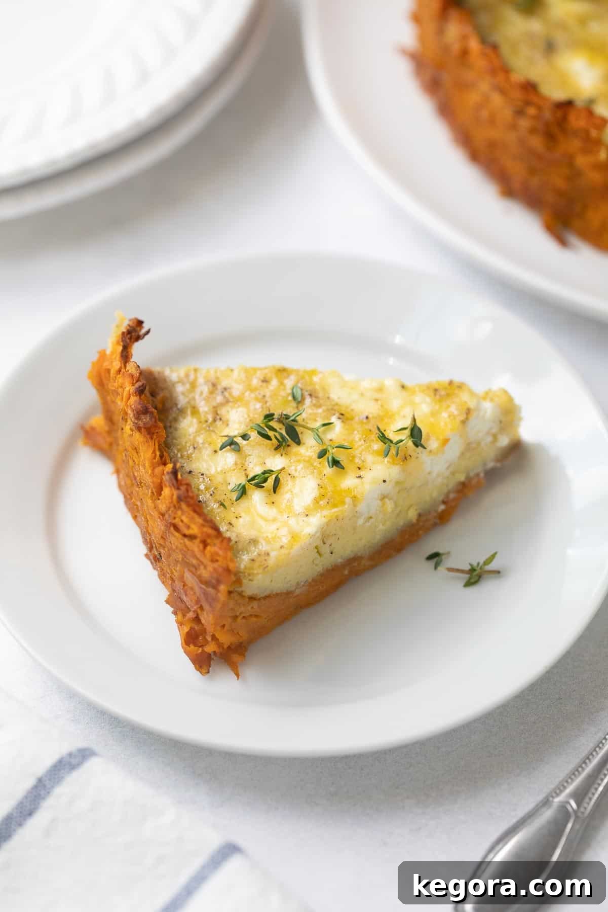 Golden Sweet Potato Quiche with Creamy Goat Cheese and Leeks 7 A slice of sweet potato crust quiche on a white plate, garnished with thyme and served with a fork and white napkin