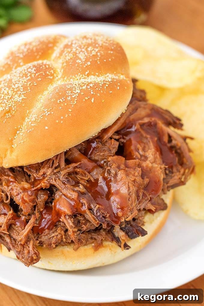 Instant Pot BBQ Pot Roast 2 Barbecue Beef on a hamburger bun with potato chips on a white plate, showcasing a delicious, shredded beef sandwich.