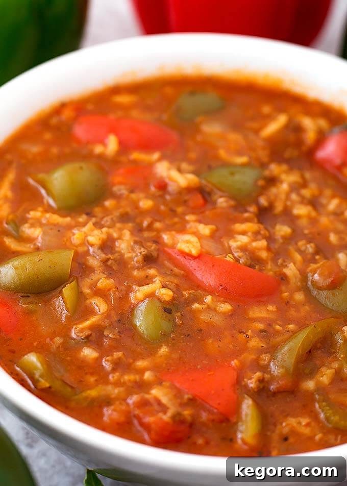Close-up view of Stuffed Pepper Soup in a pristine white bowl with a spoon, garnished, and fresh green and red bell peppers visible in the soft background