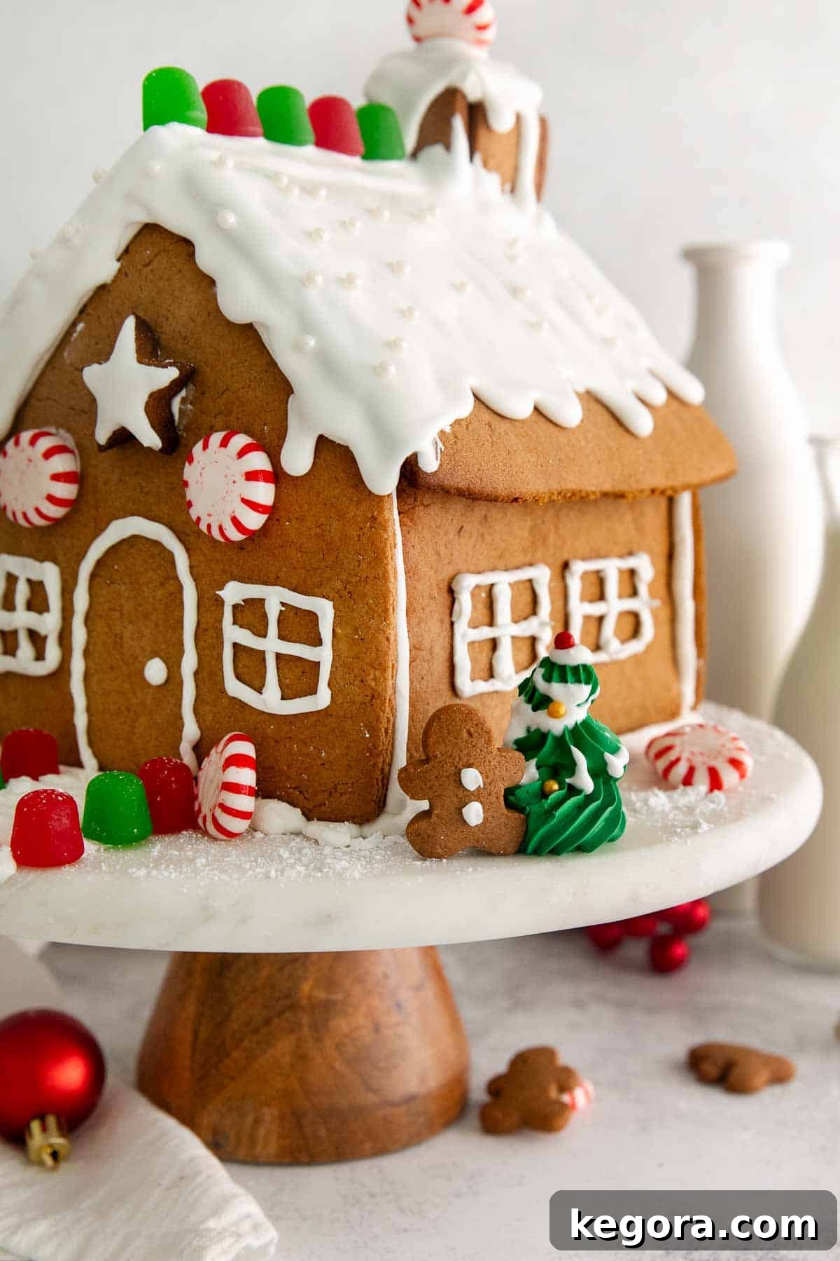 Gluten-Free Gingerbread Wonderland 17 A beautifully decorated gluten-free gingerbread house on a cake platter, surrounded by holiday ornaments and vases, ready to be enjoyed or displayed.