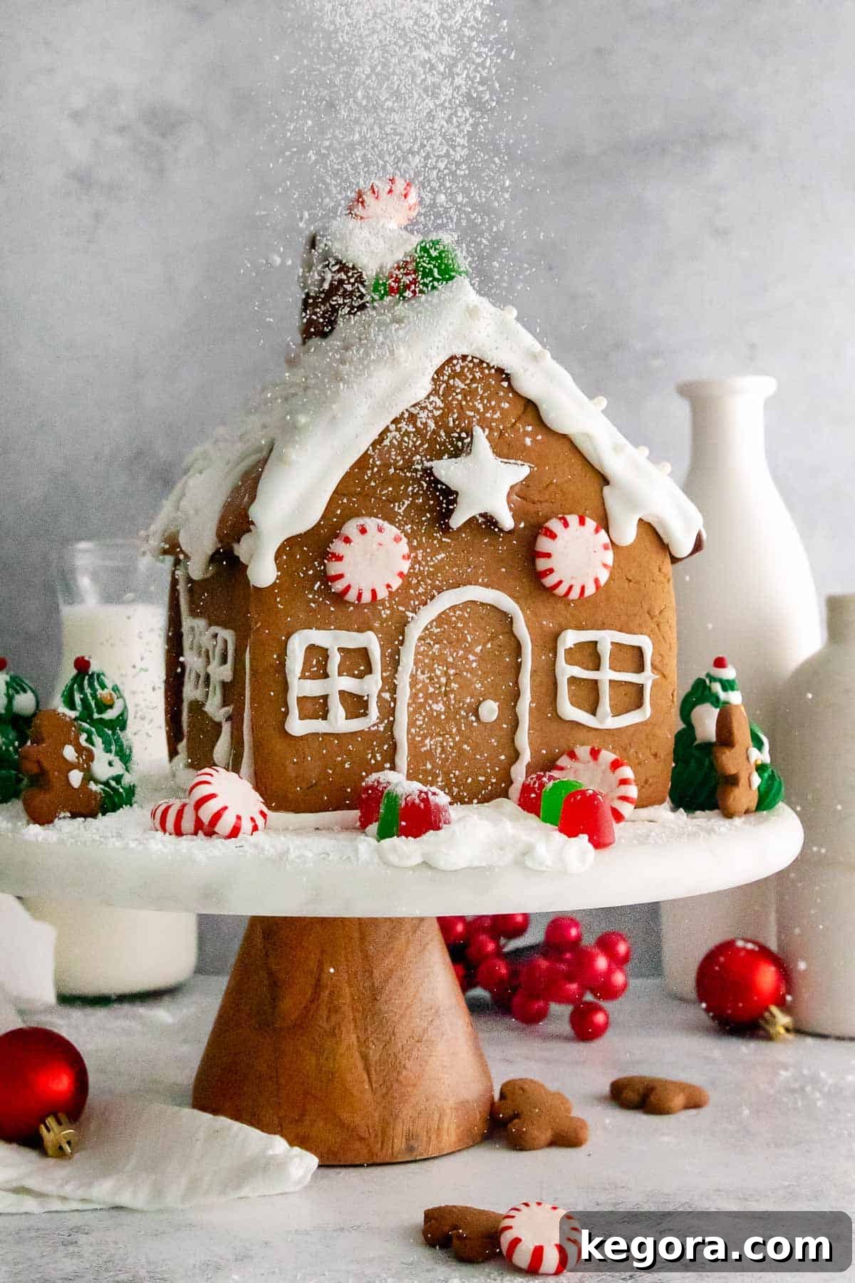 Beautifully decorated gluten-free gingerbread house on a cake platter with festive holiday ornaments surrounding it