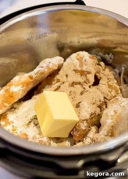 Chicken breasts and a cube of butter resting on a bed of pepperoncinis and seasoning in an Instant Pot prior to cooking.