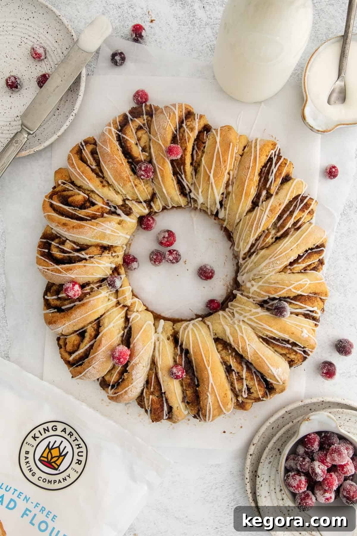Irresistible Gluten-Free Cinnamon Swirl Wreath 3 A beautifully arranged gluten-free cinnamon roll wreath, generously topped with a sweet glaze and festive sugared cranberries, elegantly presented on parchment paper.