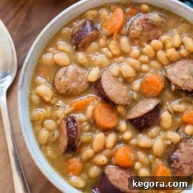 Sausage and White beans in a gray bowl next to a silver spoon on a wooden board