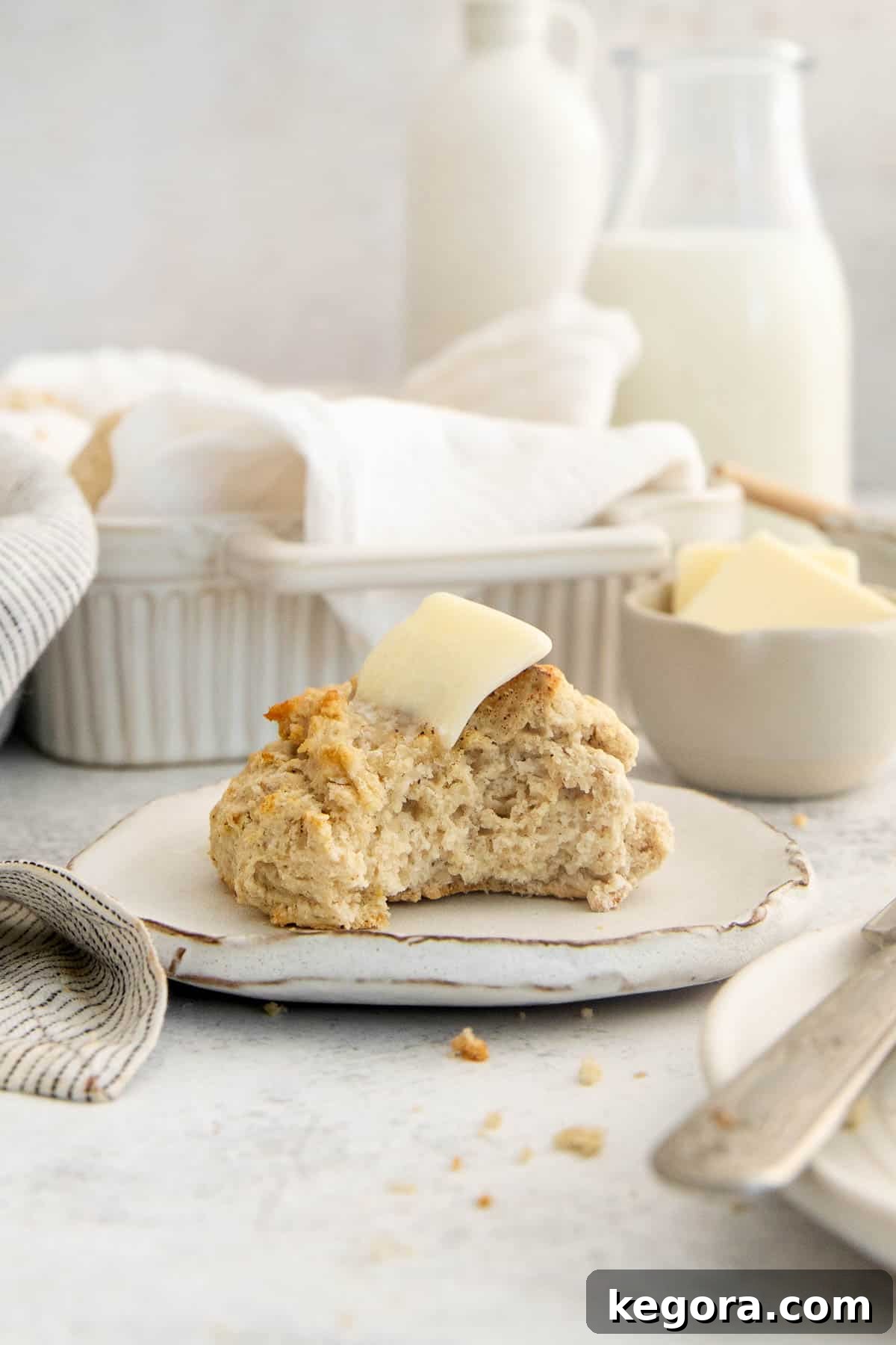 A single gluten-free drop biscuit on a white plate with one bite taken out, topped with melted butter, showcasing its soft interior.