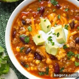 Taco Soup garnished with cubed avocado in a white bowl next to silver bowl on green mat
