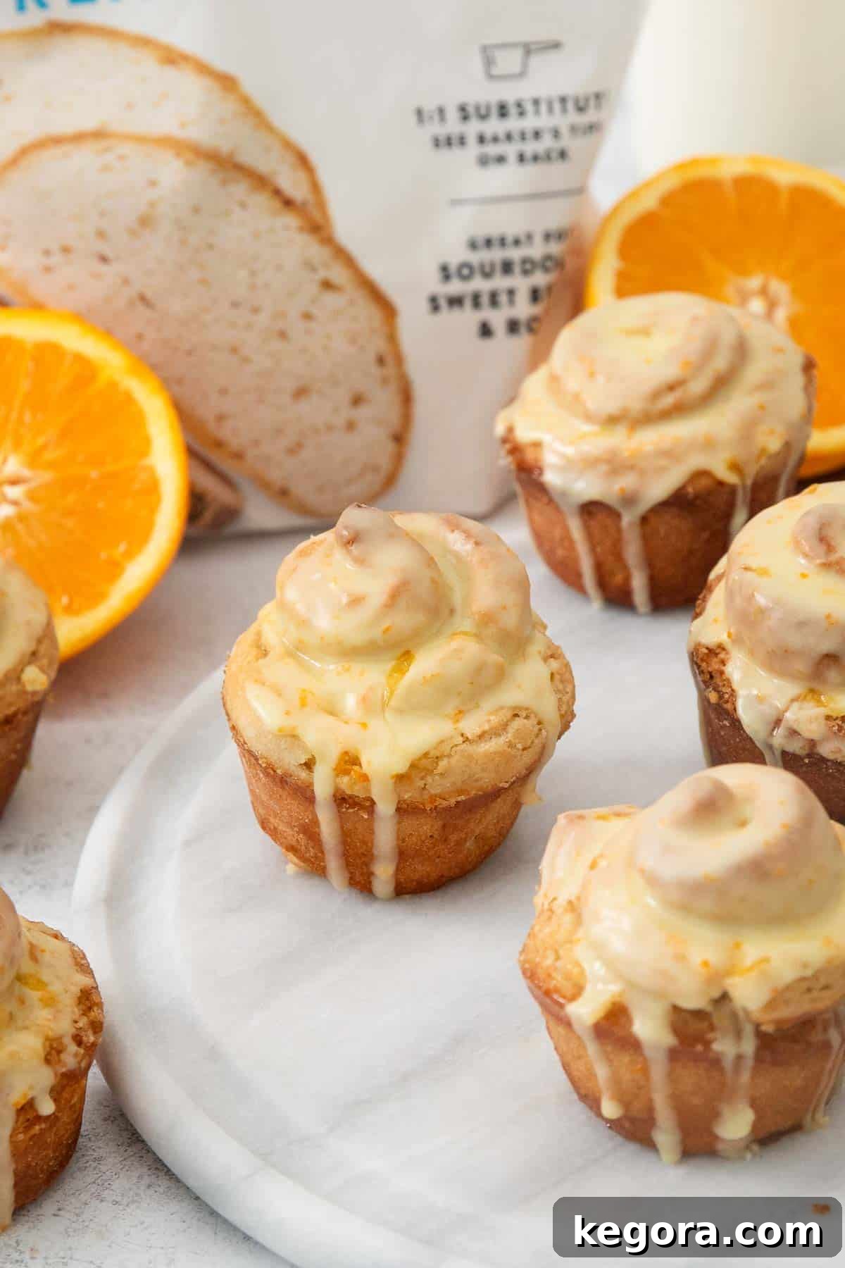 Zesty Gluten-Free Orange Buns 10 A beautiful arrangement of freshly baked and glazed gluten-free orange rolls on a platter, with whole oranges, flour, and baking ingredients artfully blurred in the background, creating a warm and inviting scene.