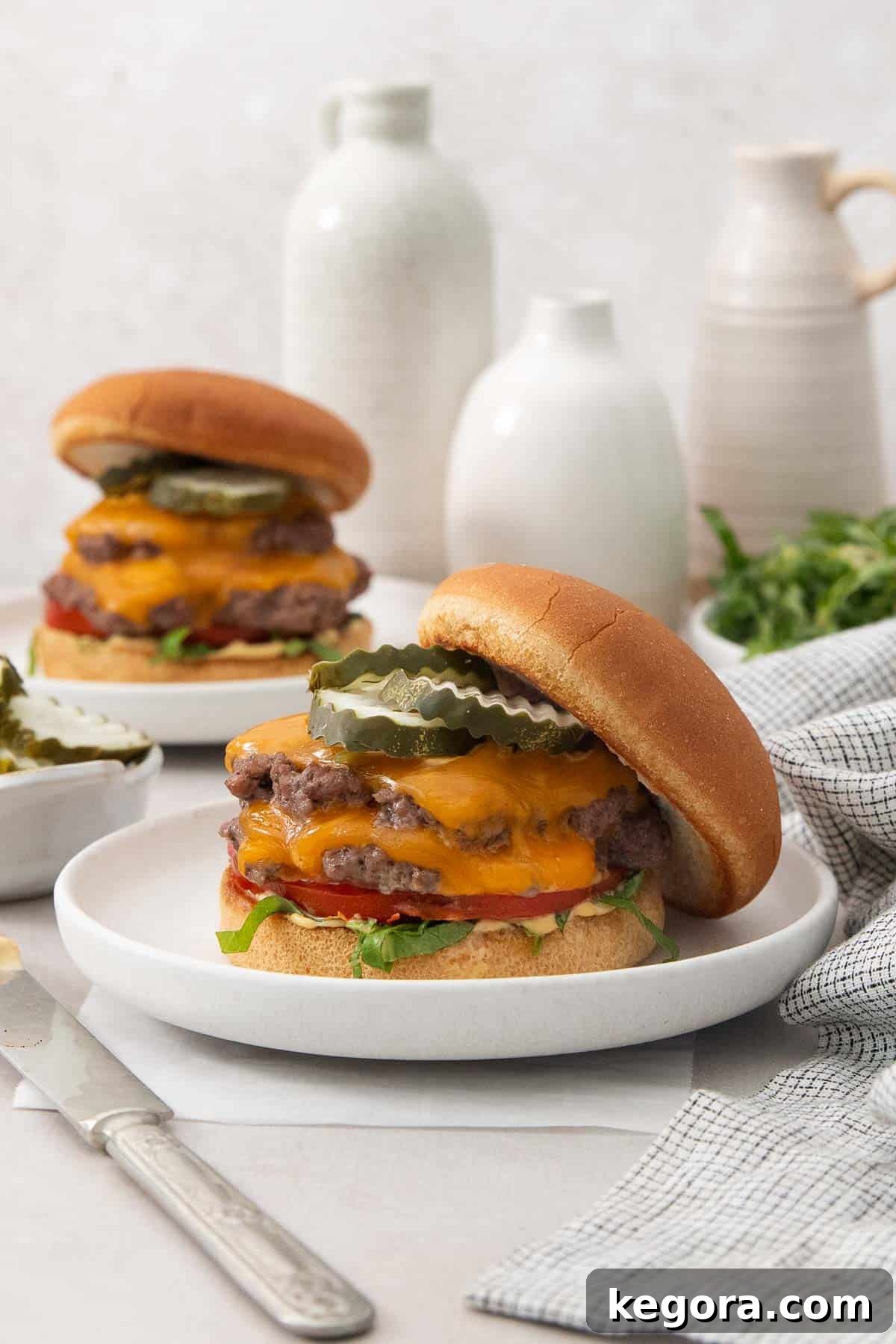 Two perfectly cooked gluten-free smashburger patties, generously topped with melted cheese, fresh lettuce, sliced tomatoes, pickles, and a dollop of our signature sauce, all nestled in toasted buns on a white surface, ready to be enjoyed.