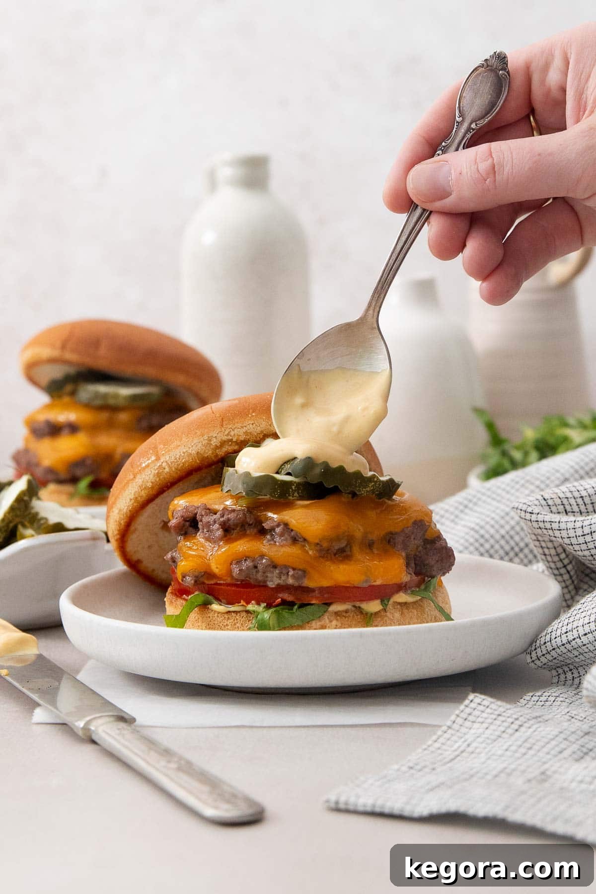 A close-up of a homemade gluten-free smashburger, topped with melted cheese, crisp lettuce, juicy tomato, and pickles, with a spoon drizzling extra smashburger sauce over the patty. The toasted bun and vibrant ingredients highlight its deliciousness.