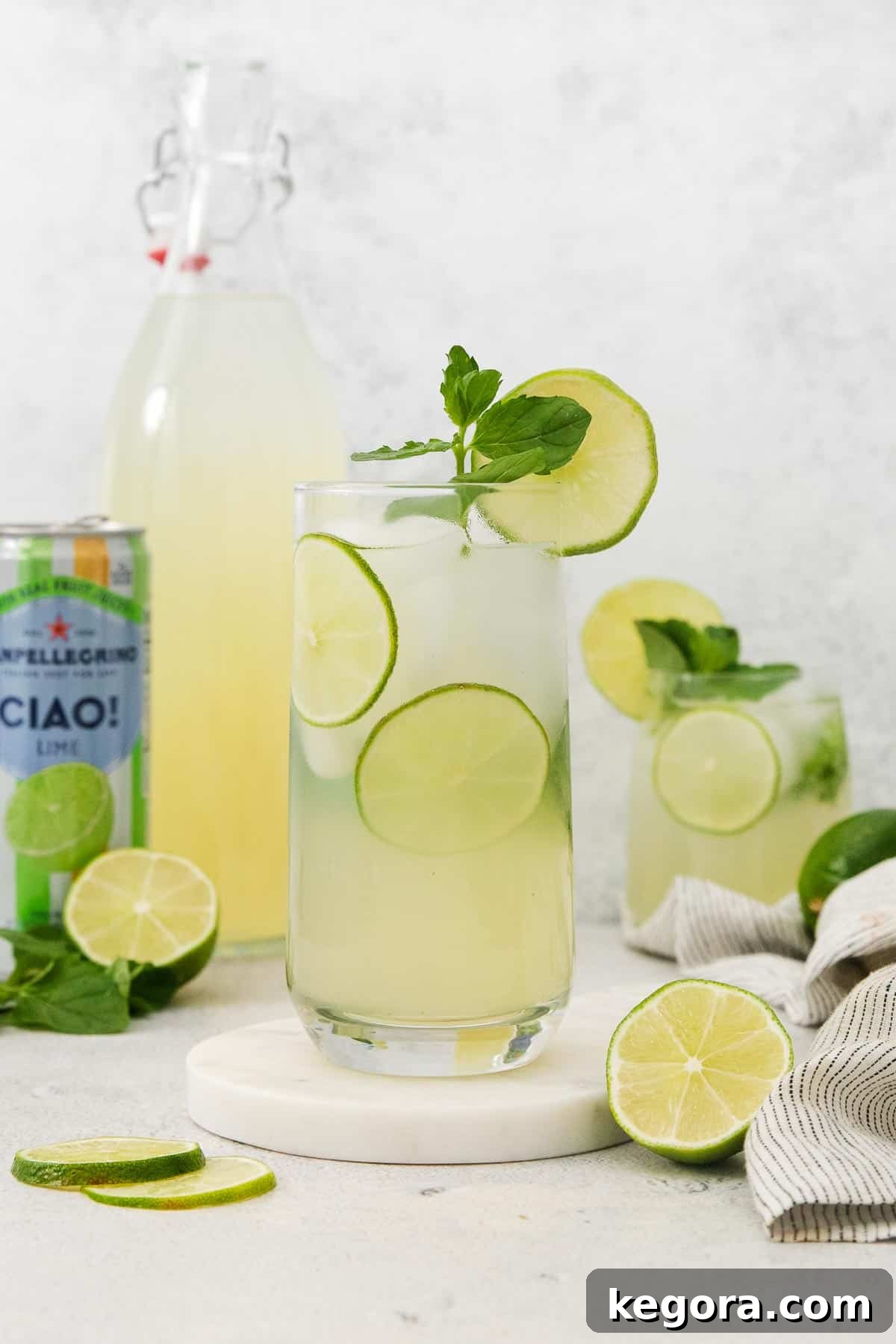 Mint Lime Cooler 3 A refreshing glass of homemade mint limeade on a countertop, with a bottle of mint limeade concentrate and sparkling lime water in the background, garnished with fresh mint.
