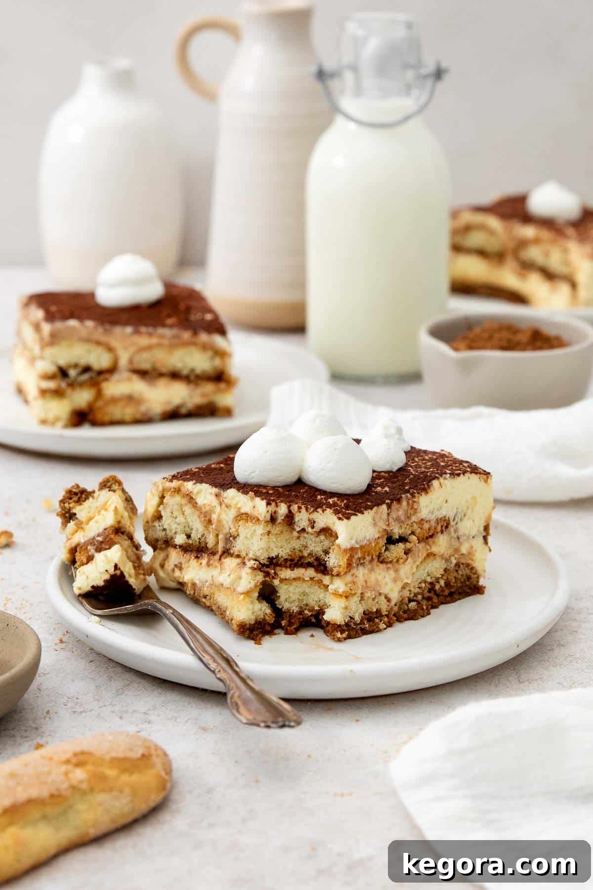 A perfectly portioned slice of gluten-free tiramisu, showcasing its distinct layers, on a pristine white plate. A fork is poised to take a bite, highlighting the dessert's creamy texture.