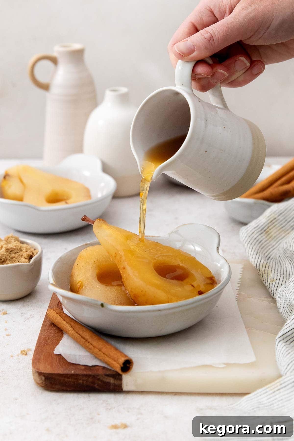 A close-up of two poached pears in a rustic dish, with a hand gently pouring a glistening apple cider reduction over them.